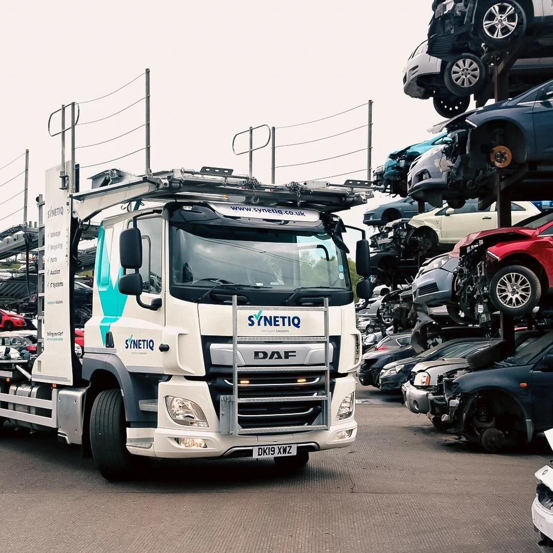 A white Synetiq truck parked in a car junkyard surrounded by stacked dismantled cars and car parts.