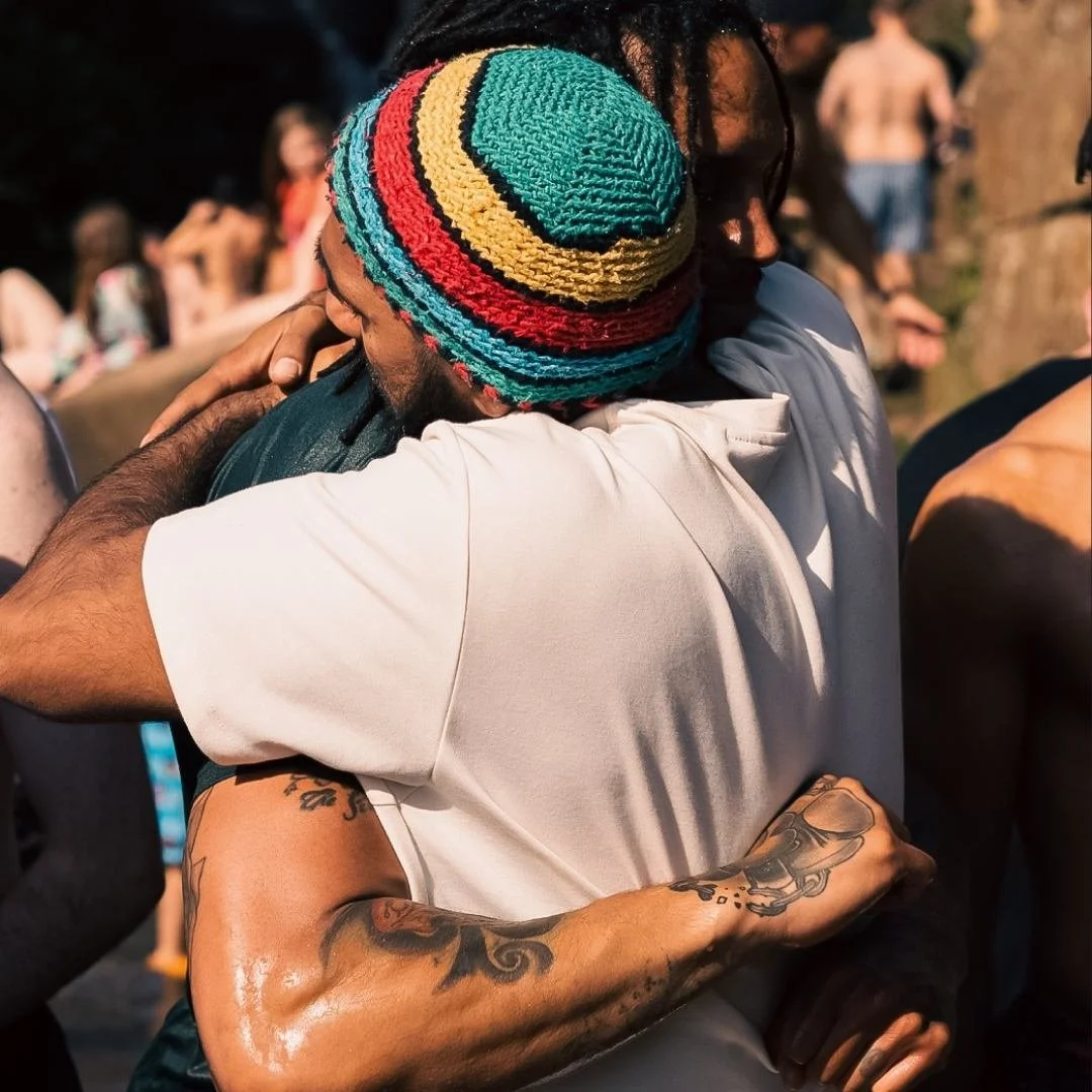 Two men hugging each other warmly outdoors, one wearing a colorful knit hat and the other in a Mandem Meetup t-shirt, with blurred people in the background.