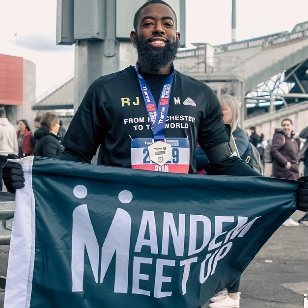 A man with a beard smiling and holding a dark blue banner that says "Mandem Meetup" at Manchester marathon. He is wearing a black shirt with text "mandem meetup x UNO", with people and a stadium structure in the background.