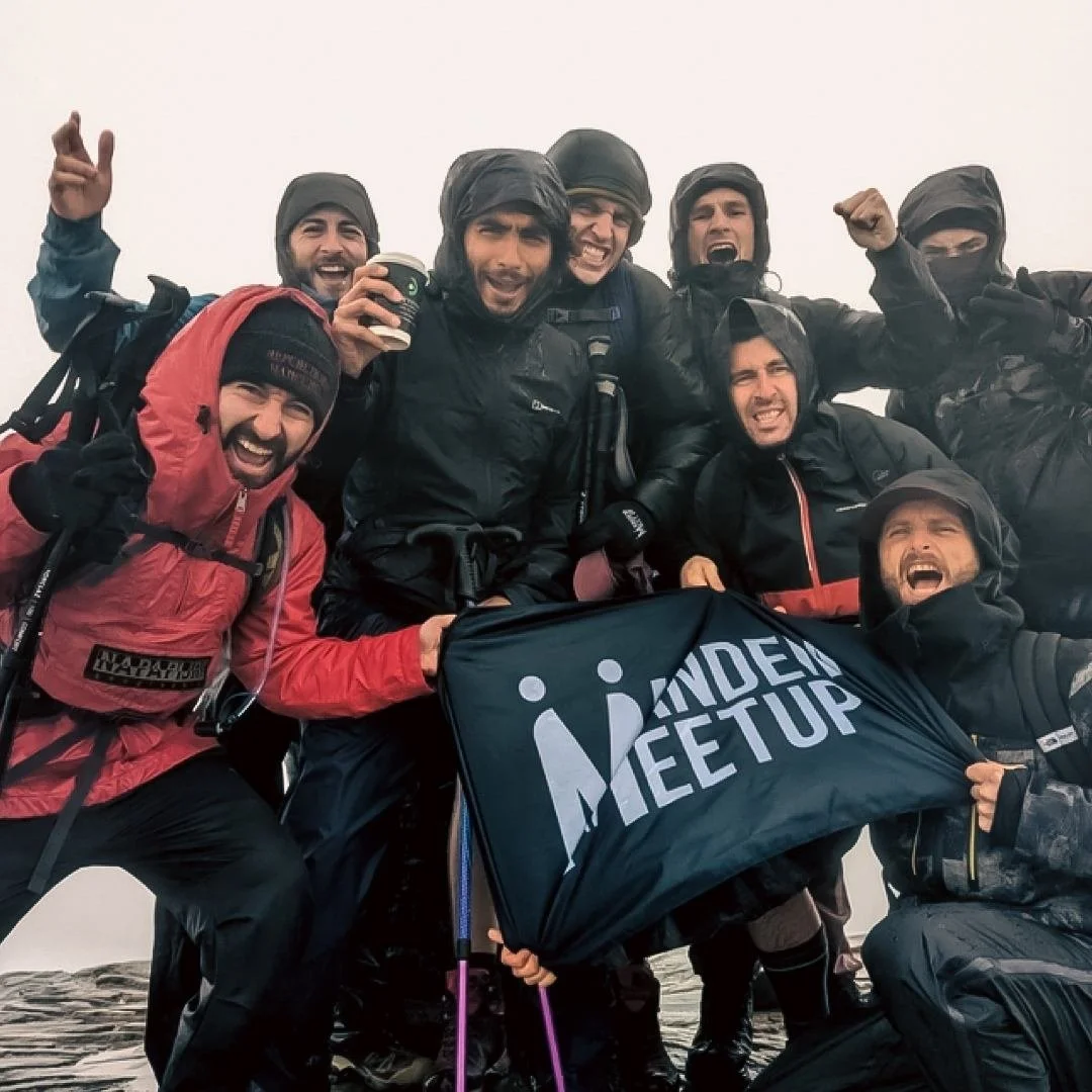 Group of eight men in outdoor gear, celebrating and smiling, holding a black 'Mandem Meetup' flag, on a foggy mountain summit.