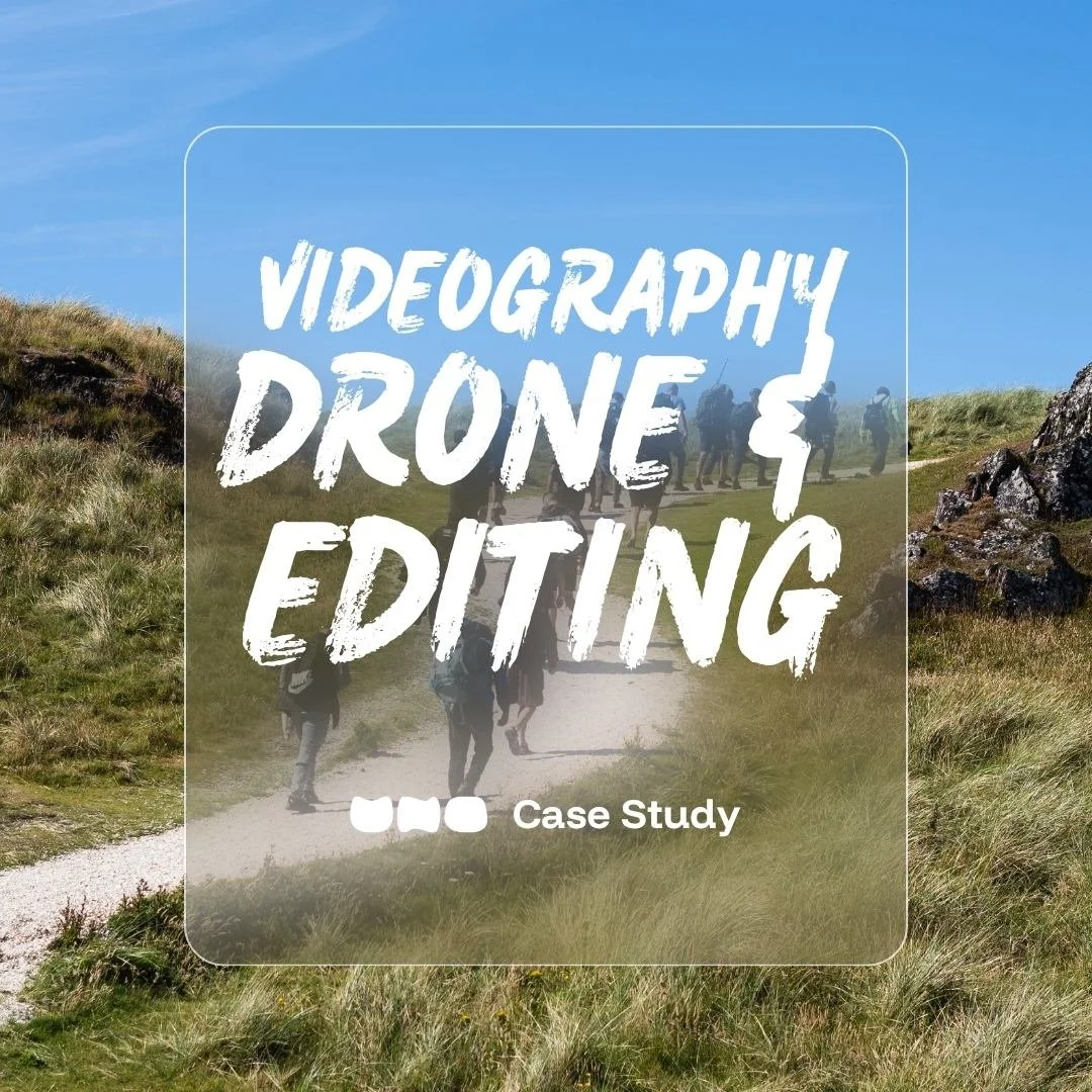 Drone shot of group of hikers walking on a dirt trail through a grassy, hilly landscape under a blue sky, some men Mandem Meetup tshirts; text overlaid stating "Videography, Drone & Editing by UNO"