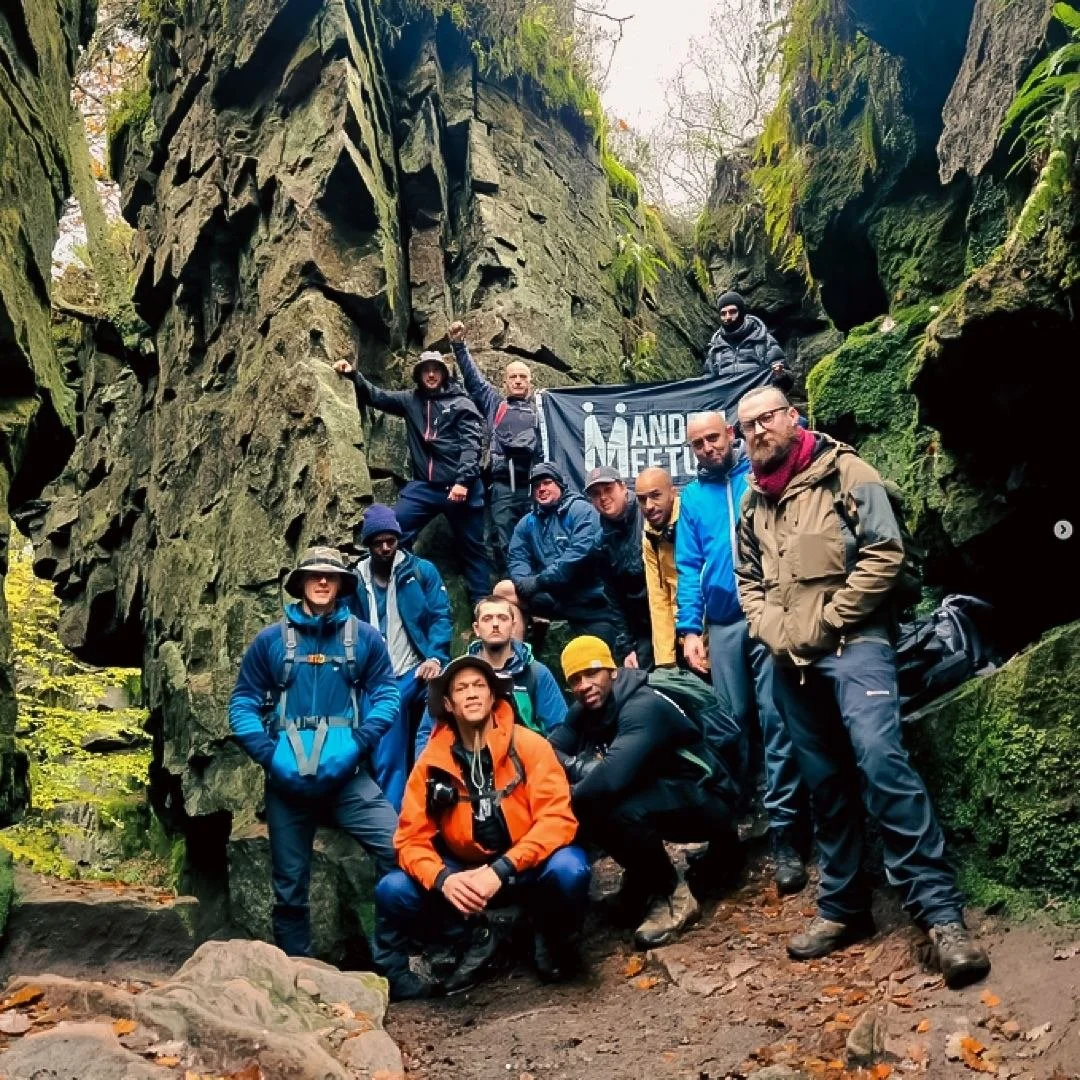 Drone shot of a group of hikers in outdoor gear posing in a rocky, moss-covered canyon with green foliage - holding a sign reading "Mandem Meetup"