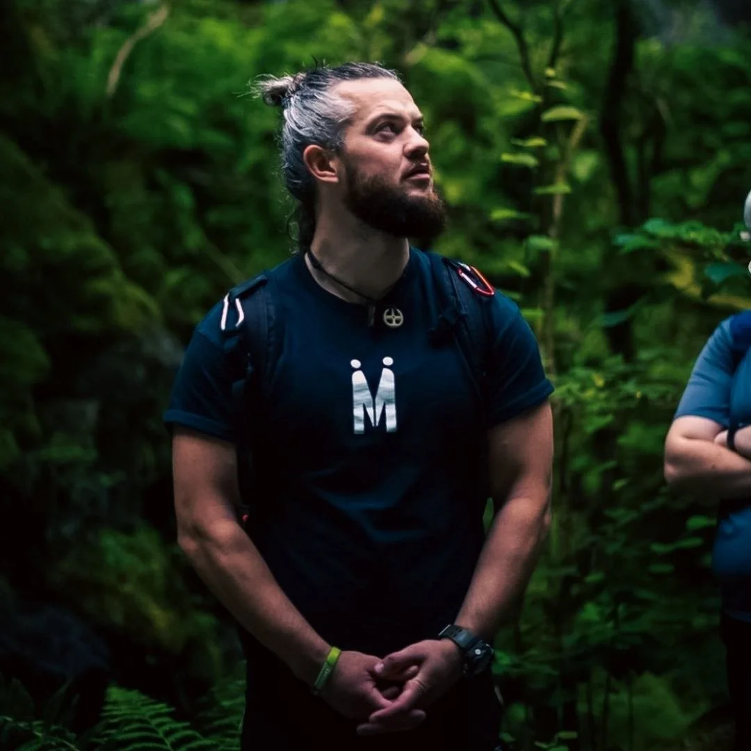 Jamie Dennis, CEO of Mandem Meetup, with a bun hairstyle and beard standing in a dense green forest, wearing a black athletic shirt and a backpack, looking upward thoughtfully - with a videographer partially in shot.