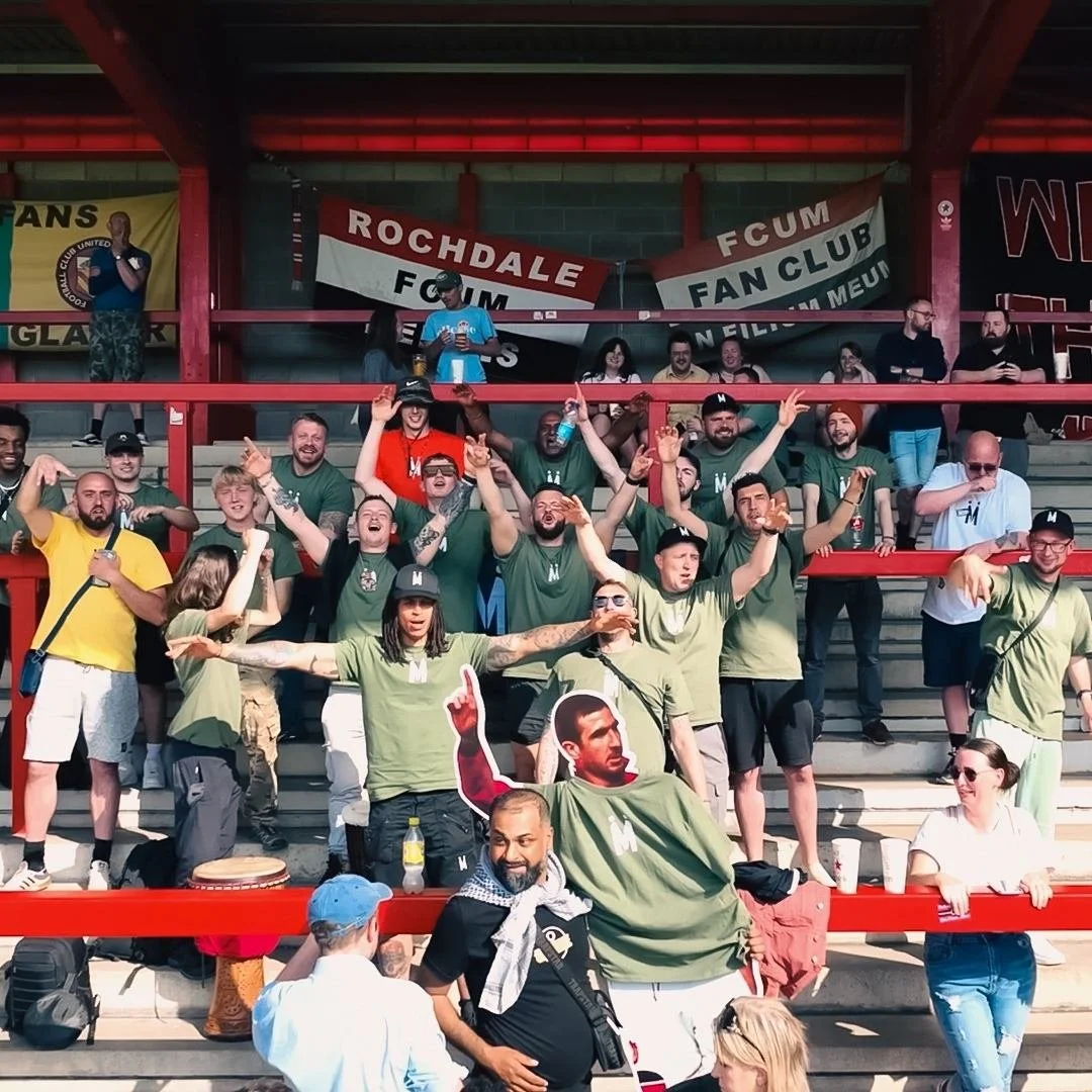 Drone shot of a group of men gathered at a football match wearing Mandem Meetup tshirts, standing on stadium stairs, cheering, and posing for a photo. Banner in the background reading "made by uno" viisible in the shot