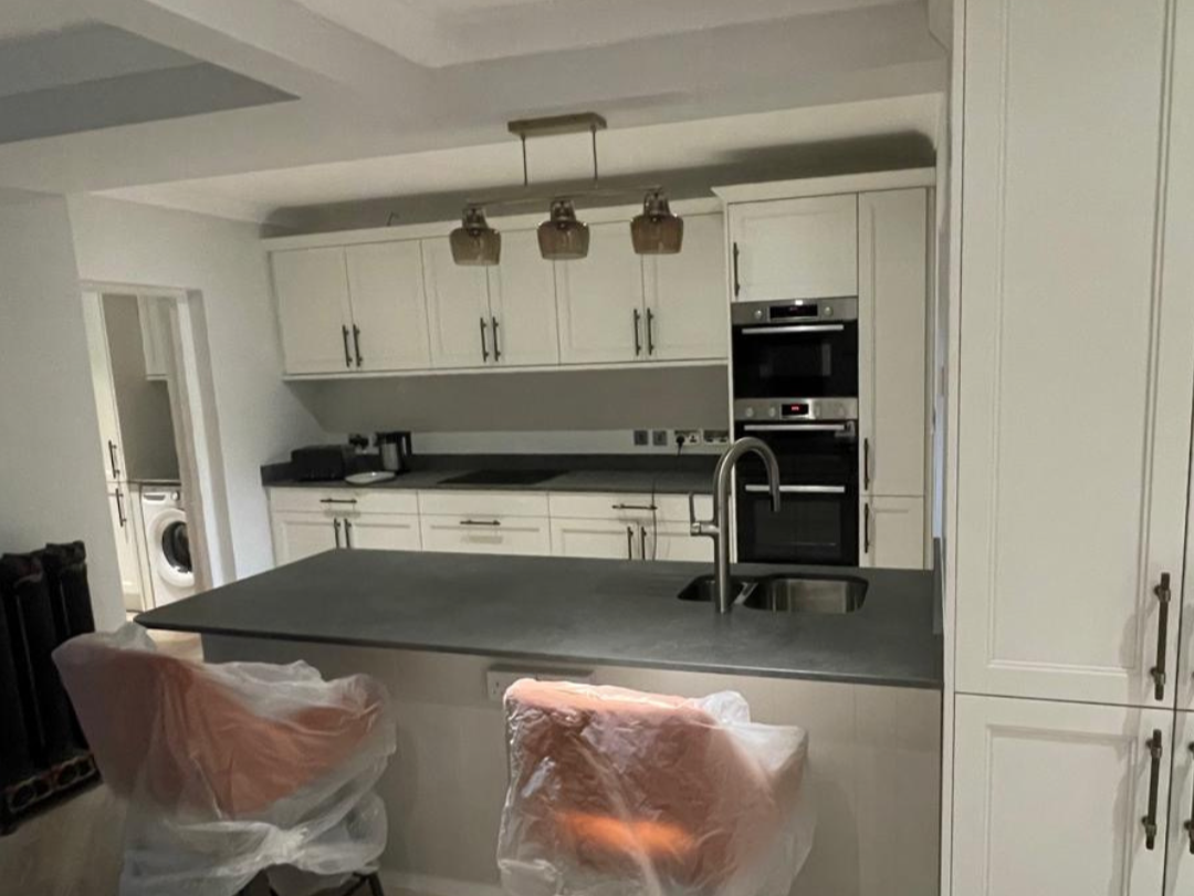 Modern kitchen with white cabinets, a gray countertop island, and built-in appliances, including an oven and microwave, with two chairs covered in plastic in the foreground.