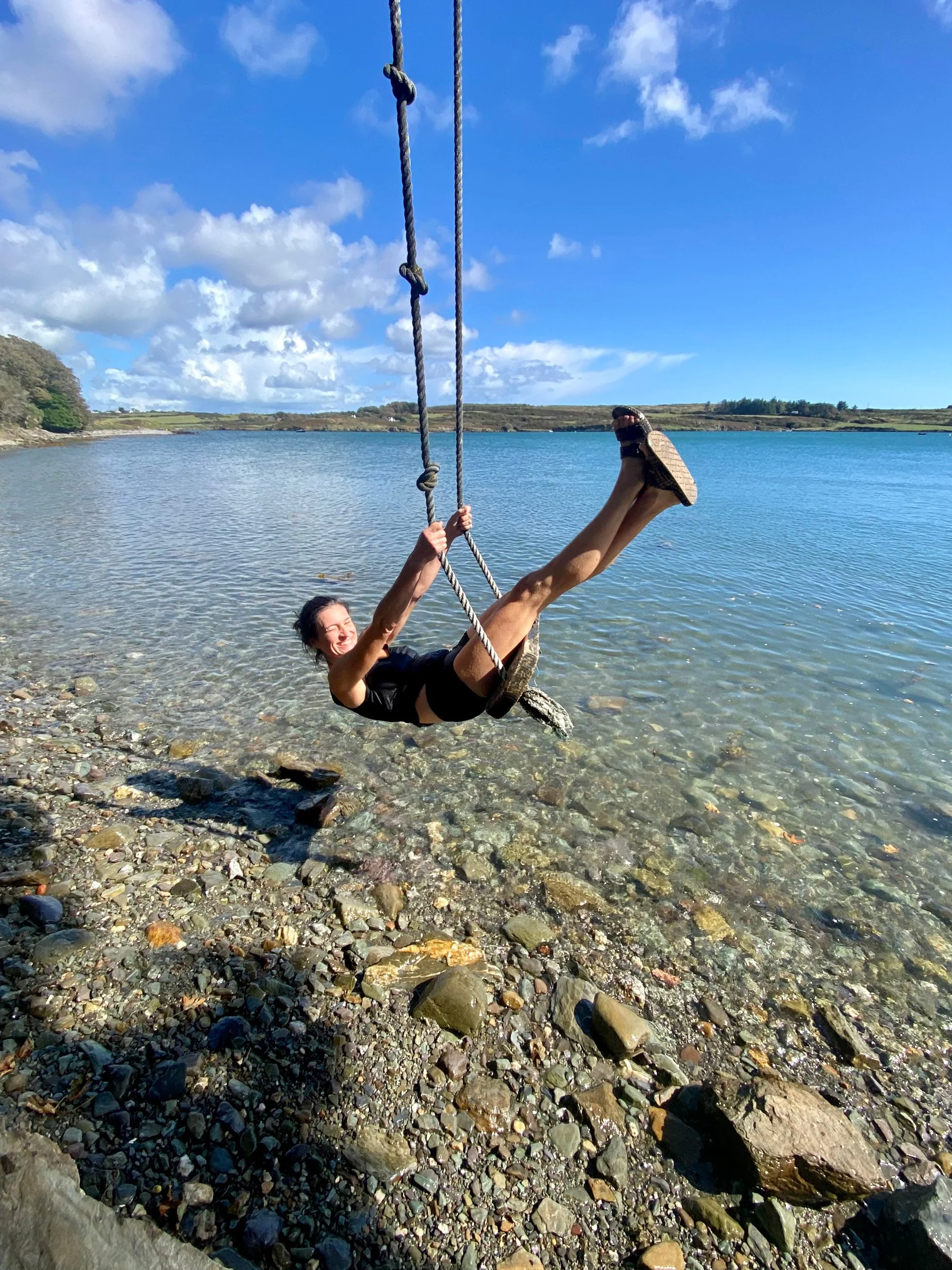 Vrouw die lachend op een touwschommel boven helder water schommelt in een natuurlijke omgeving.