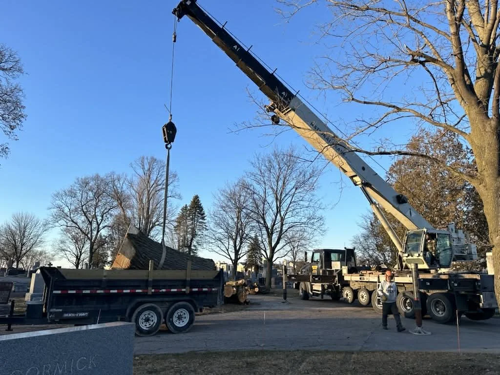 A crane lifting a large memorial stone onto a truck at an outdoor site with trees and clear sky.
