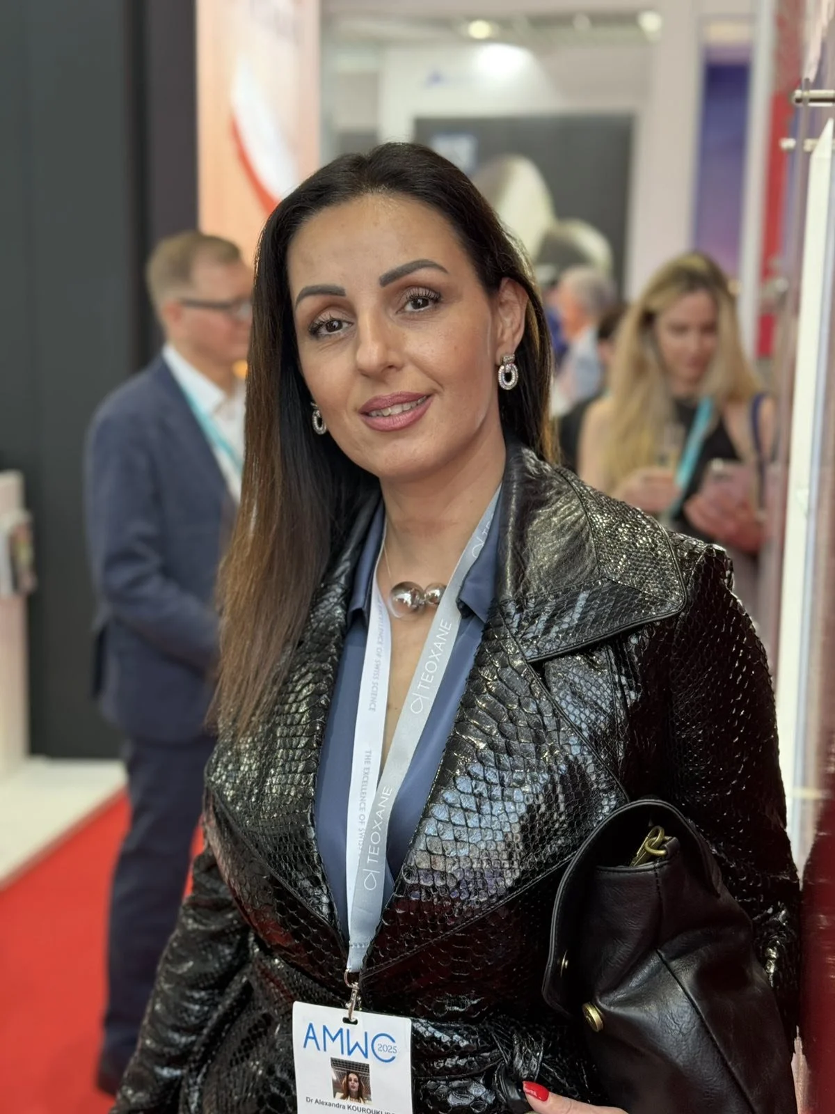 A woman with long dark hair wearing a black textured jacket, jewelry, and a lanyard at a conference. She is indoors with blurred people and booths in the background.