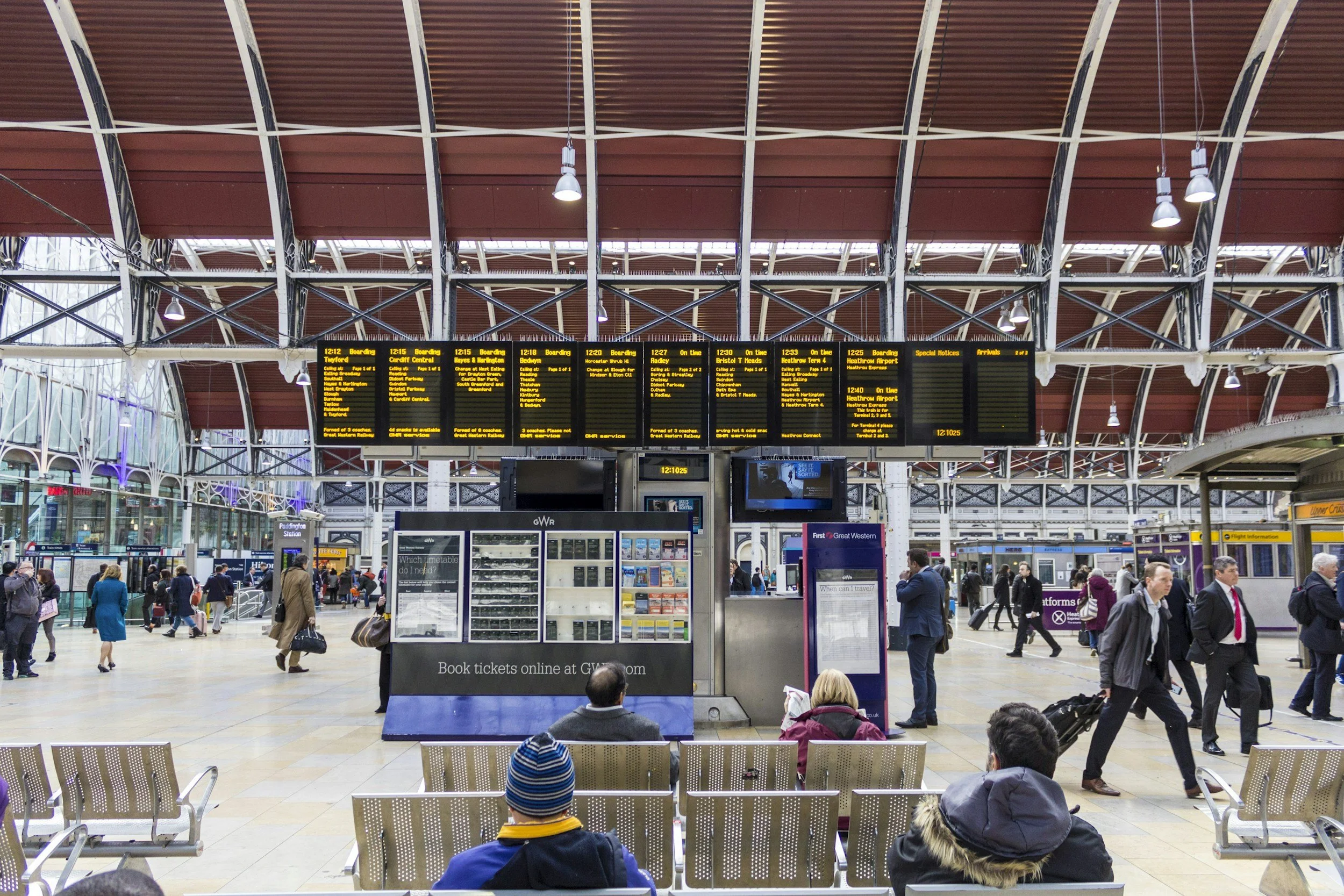 Inside a busy train station with people walking, sitting, and waiting. A large electronic departure board hangs from the ceiling displaying train information. There are benches in the foreground, and various ticket kiosks and information booths are visible.
