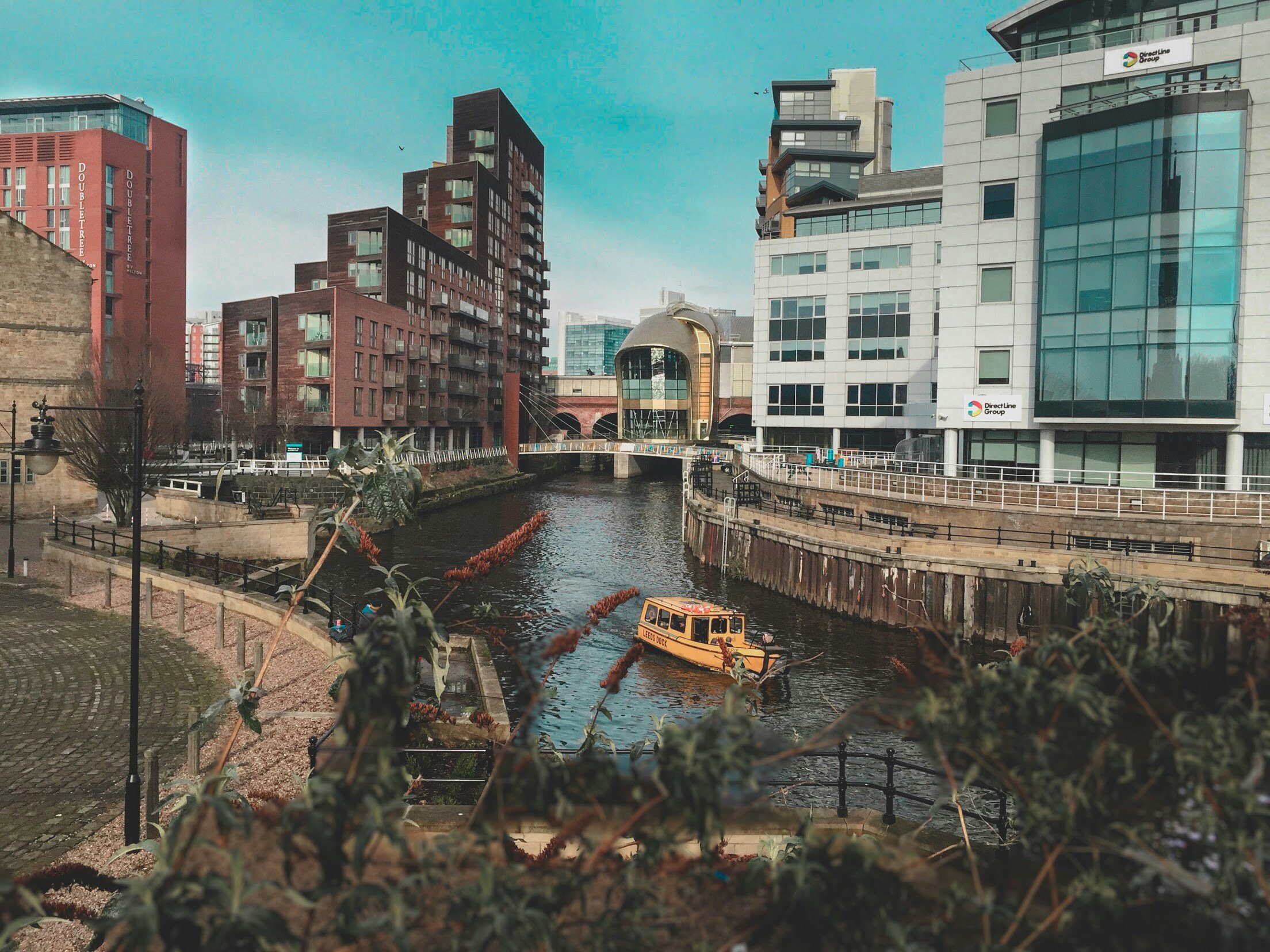 Urban scene of Leeds with canal, boat, modern multi-story buildings, and a bridge under a blue sky.