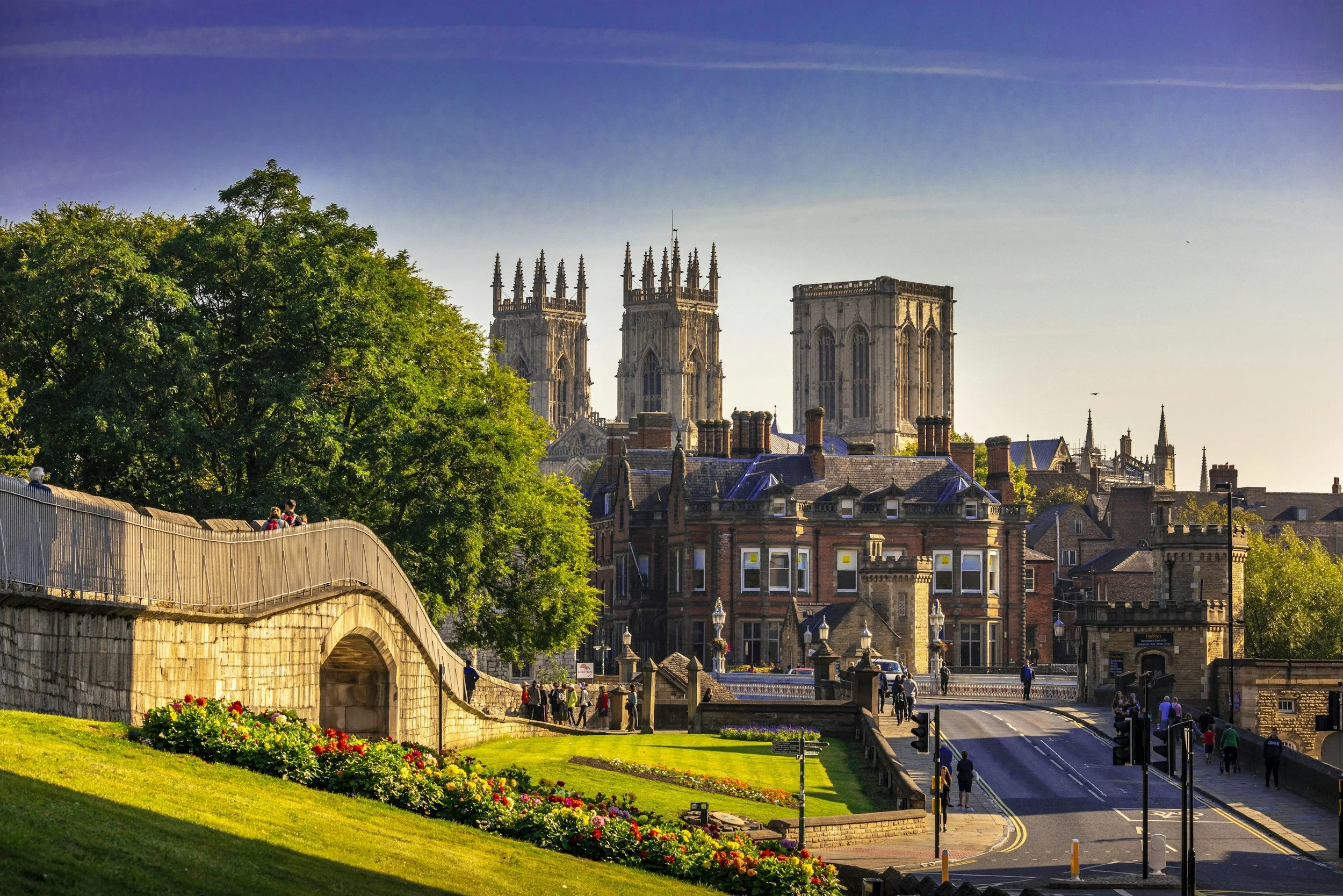 A scenic view of a York with historic buildings, including a large stone church with multiple tall, ornate towers, surrounded by smaller brick buildings, a green park with flowers, and a bridge with pedestrians under a clear sky.