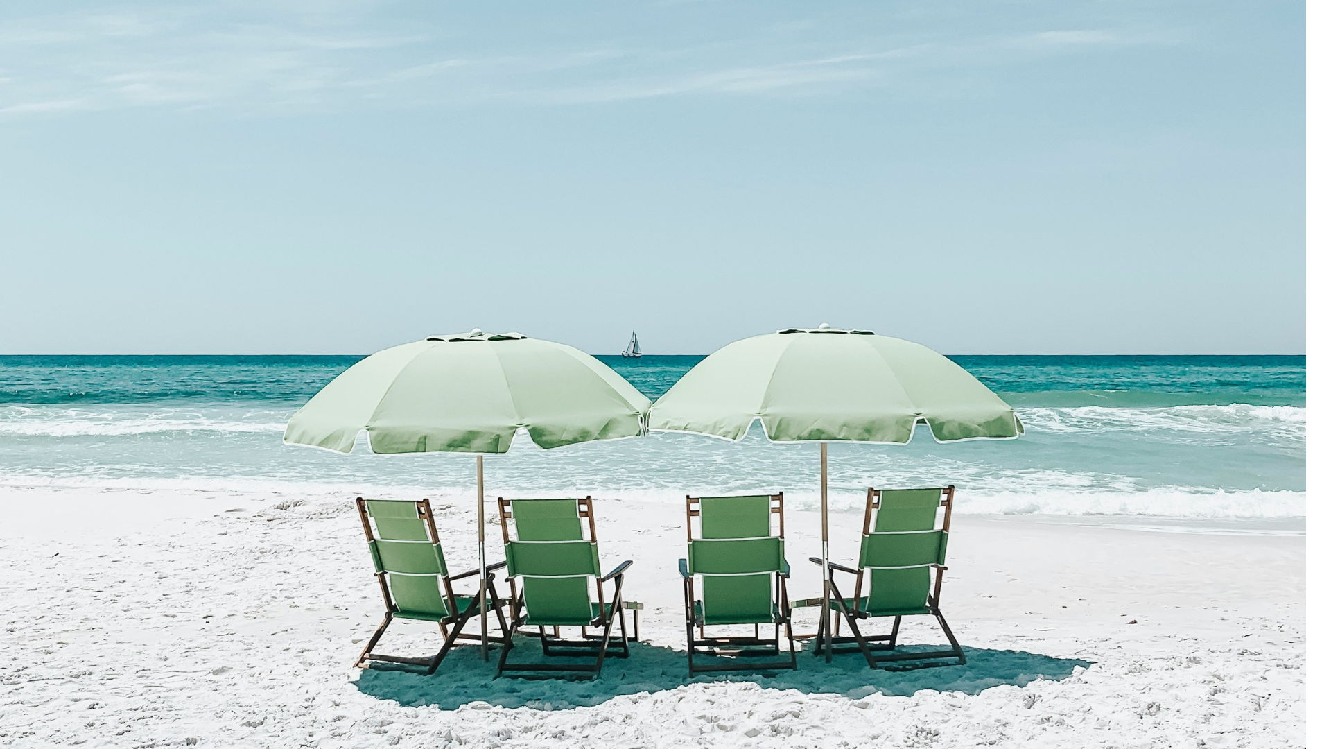 Four green beach chairs with two green umbrellas on a sandy beach with the ocean and a sailboat in the distance.