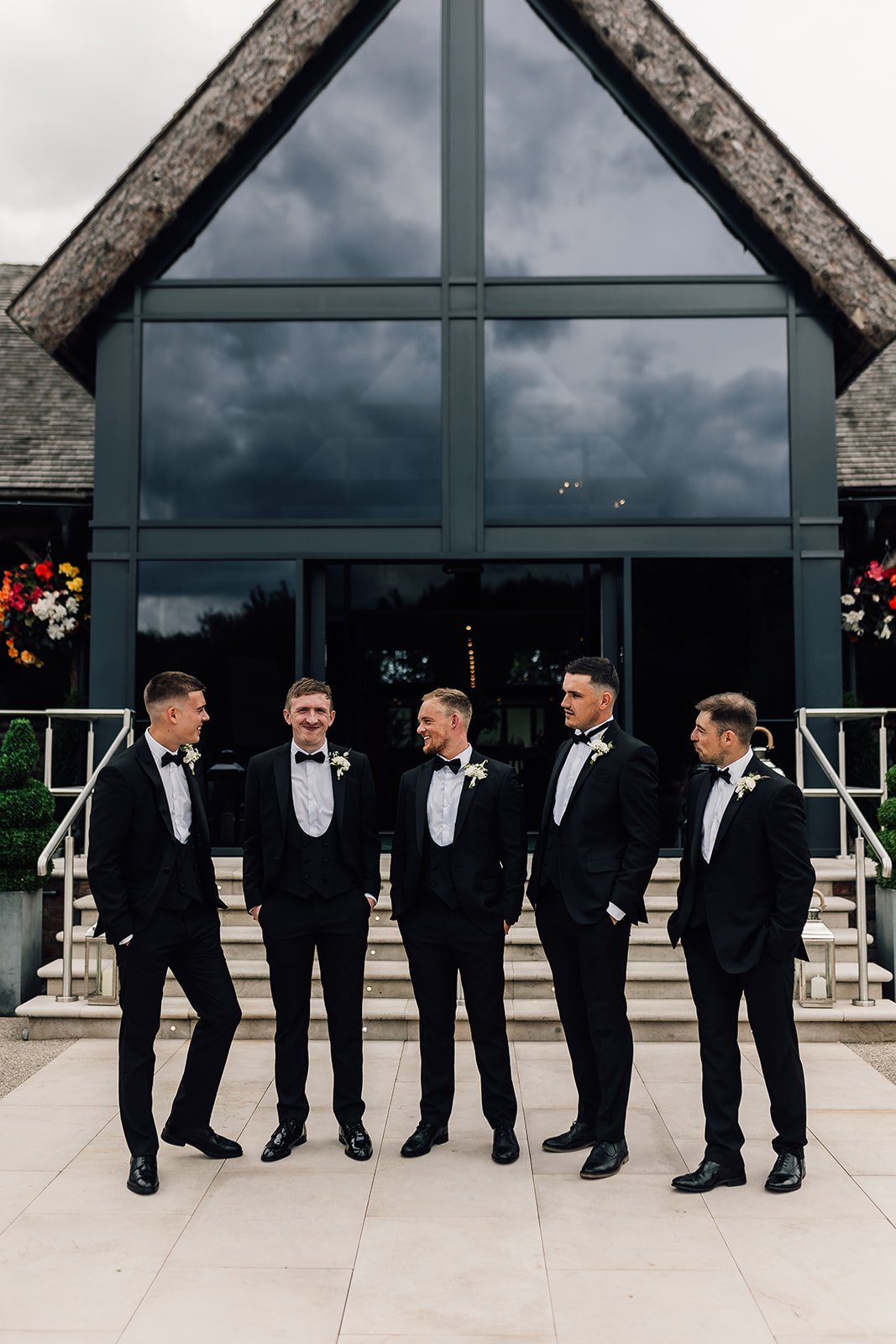 A group of six men dressed in black tuxedos and bow ties standing on steps outside a modern building with large glass windows, under a cloudy sky.