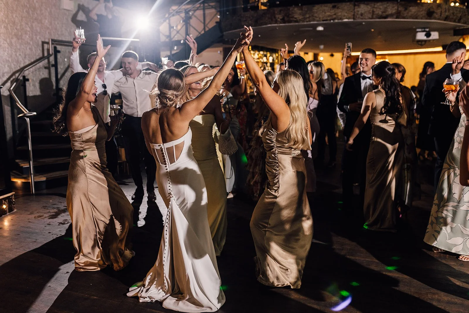People dancing and celebrating at a formal event or wedding reception, with women in elegant gowns and men in suits, illuminated by soft lighting.