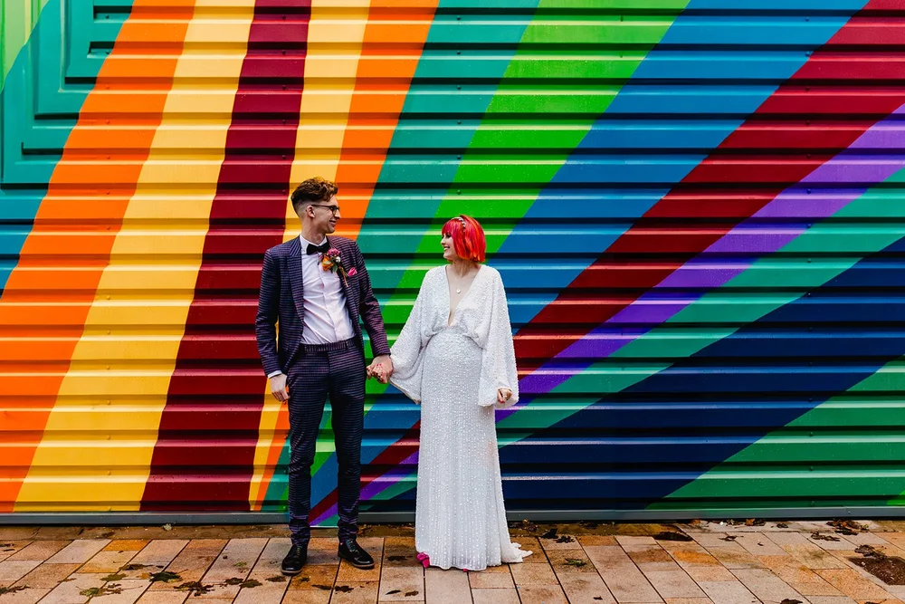 A couple in wedding attire holding hands and smiling at each other in front of a colorful rainbow mural.
