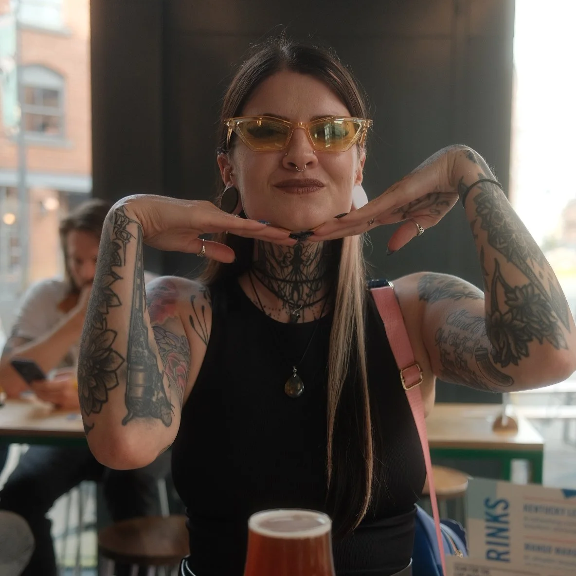 A tattooed woman with glasses and tattoos on her arms, neck, and chest, smiling and posing with her hands under her chin in a cafe or restaurant with a blurred background of other people and large windows.