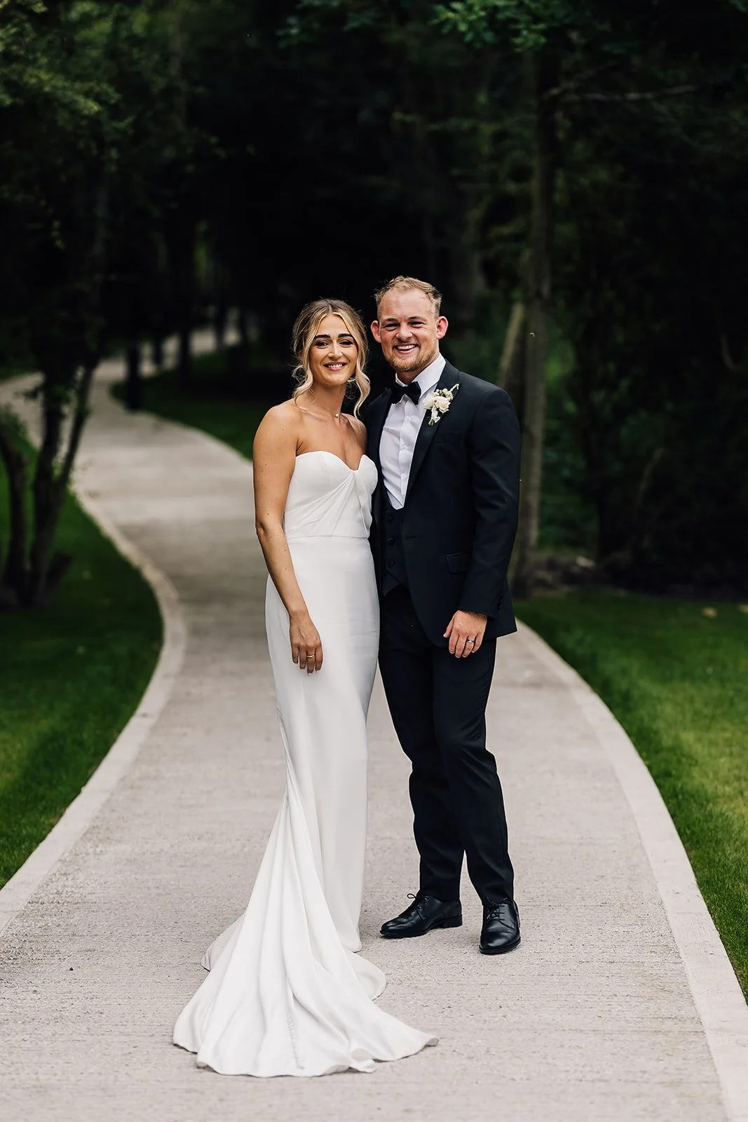 A bride and groom standing together on a paved pathway in a park with trees and greenery in the background, smiling at the camera.