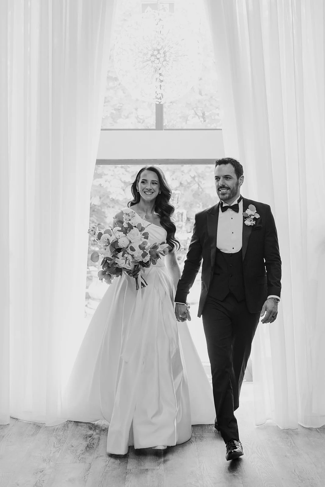Becky entering the ceremony room with her dad beneath the chandeliers