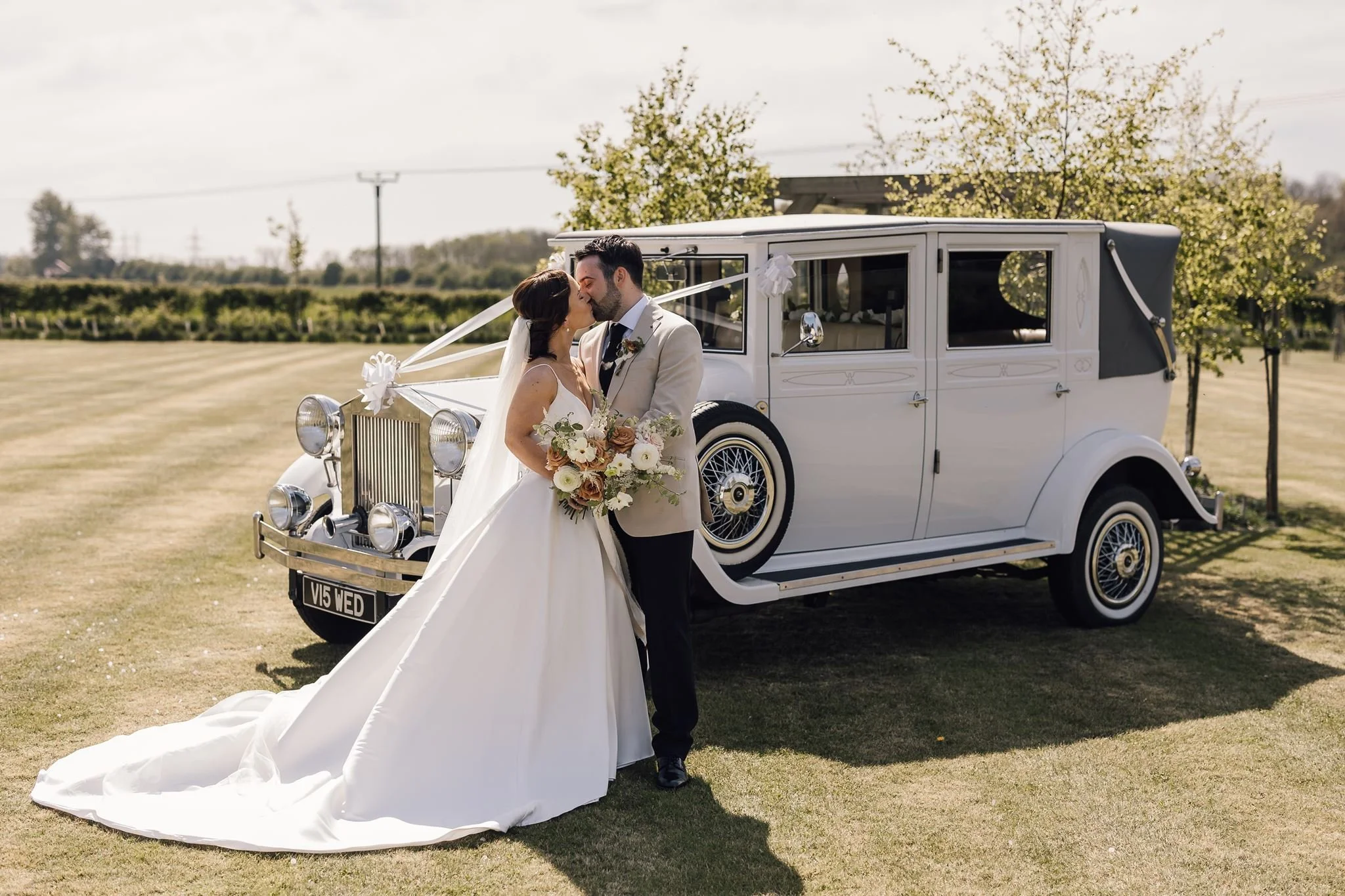 Rachel and Harry kissing beside their wedding car on the lawn