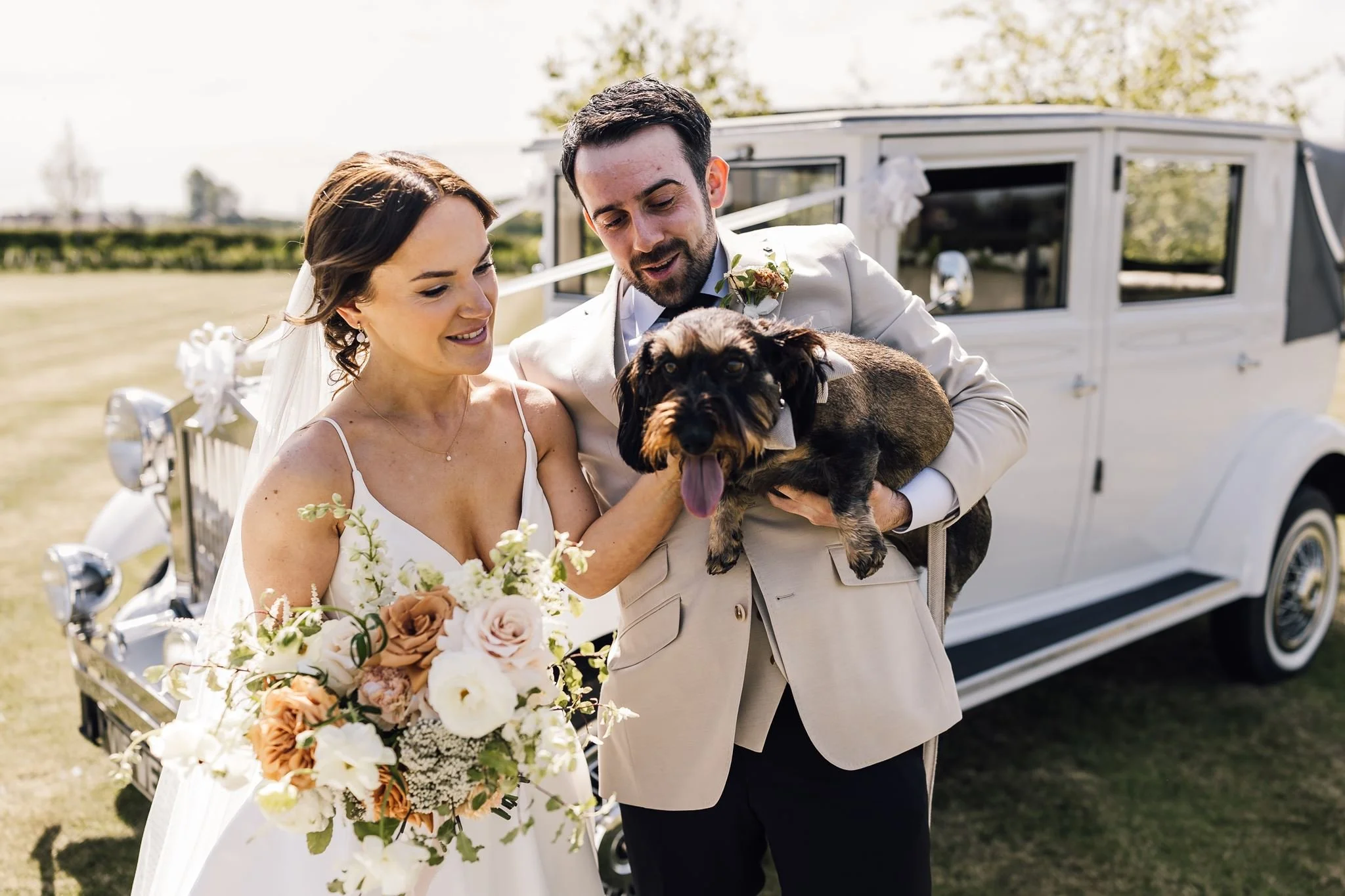 Rachel and Harry cuddling their sausage dog Dottie beside the wedding car