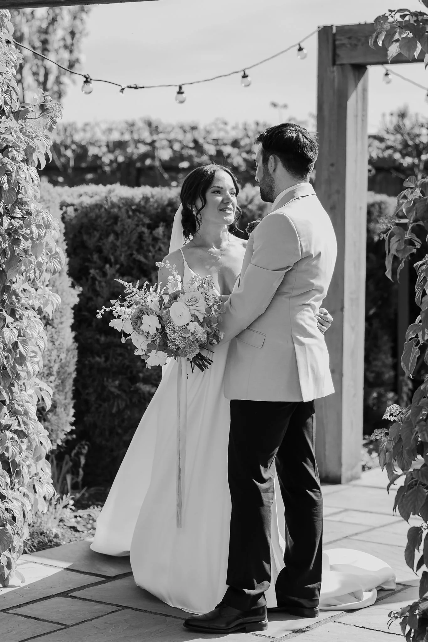 Rachel and Harry embracing beside the garden pergola, in black and white