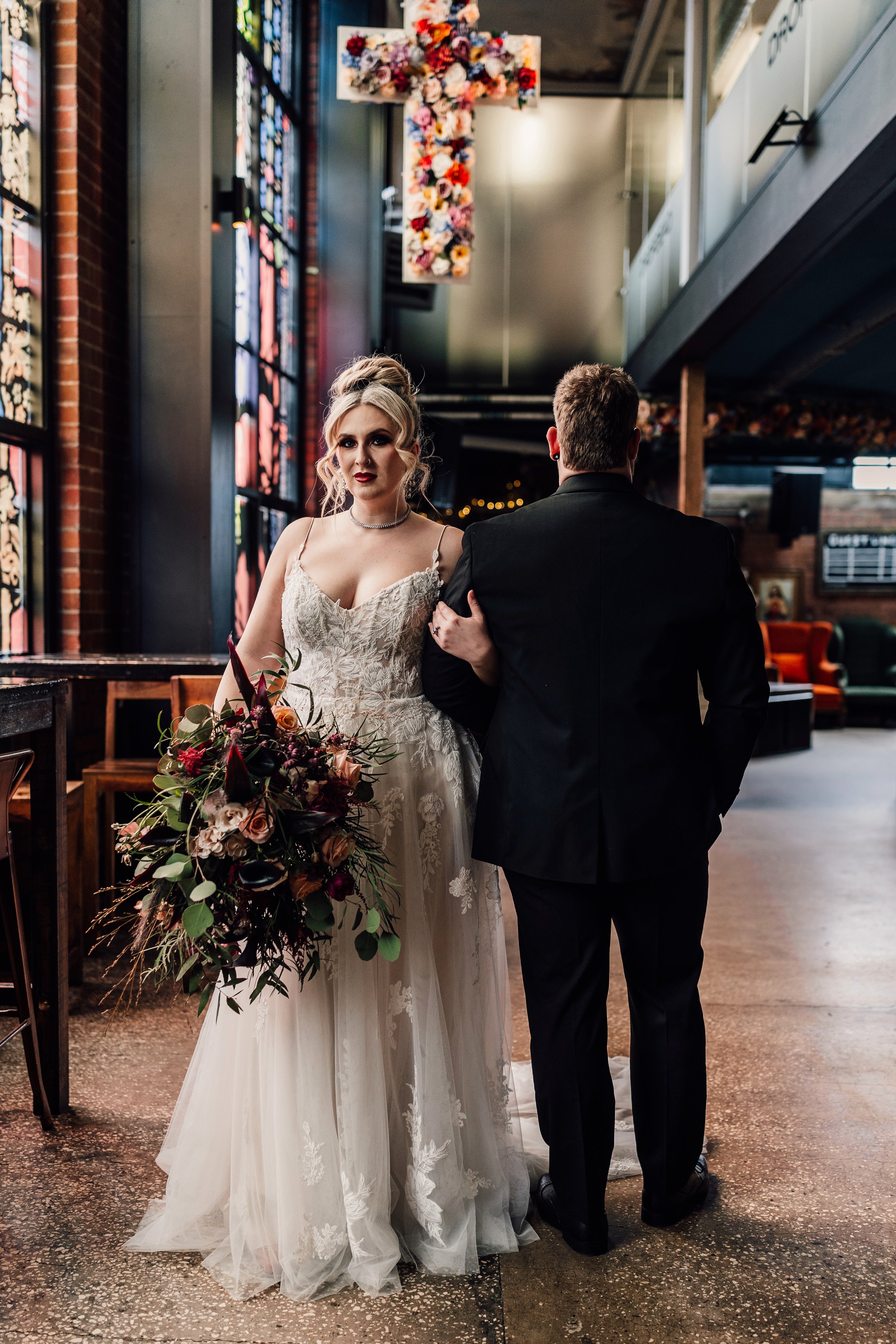 Alternative wedding couple walking through Church Temple of Fun Sheffield with stained glass windows and floral cross backdrop