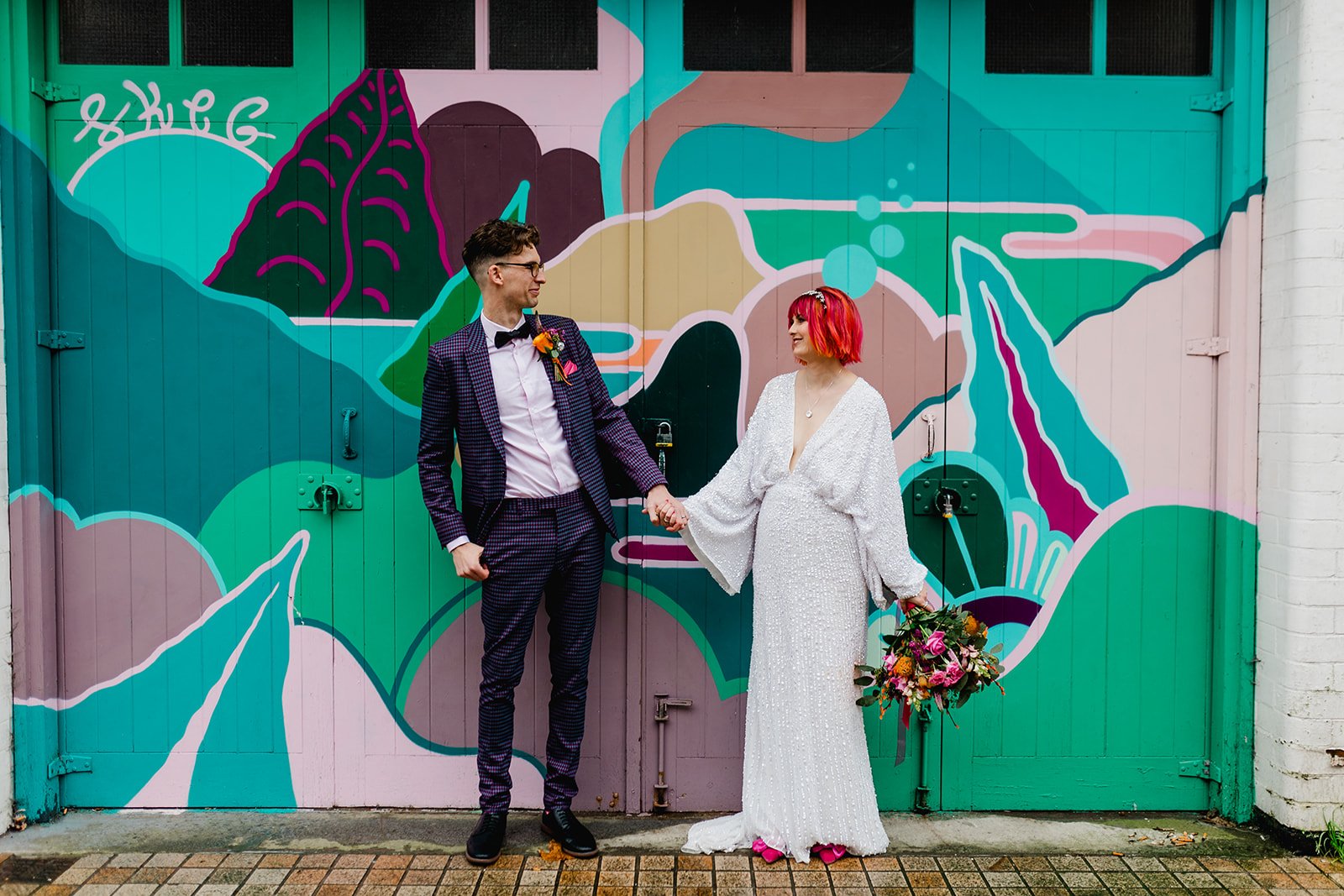 A couple holding hands in front of a colorful mural on humber street The man is dressed in a checkered suit with a bow tie, and the woman wears a white, beaded wedding gown and red hair, holding a bouquet of flowers.