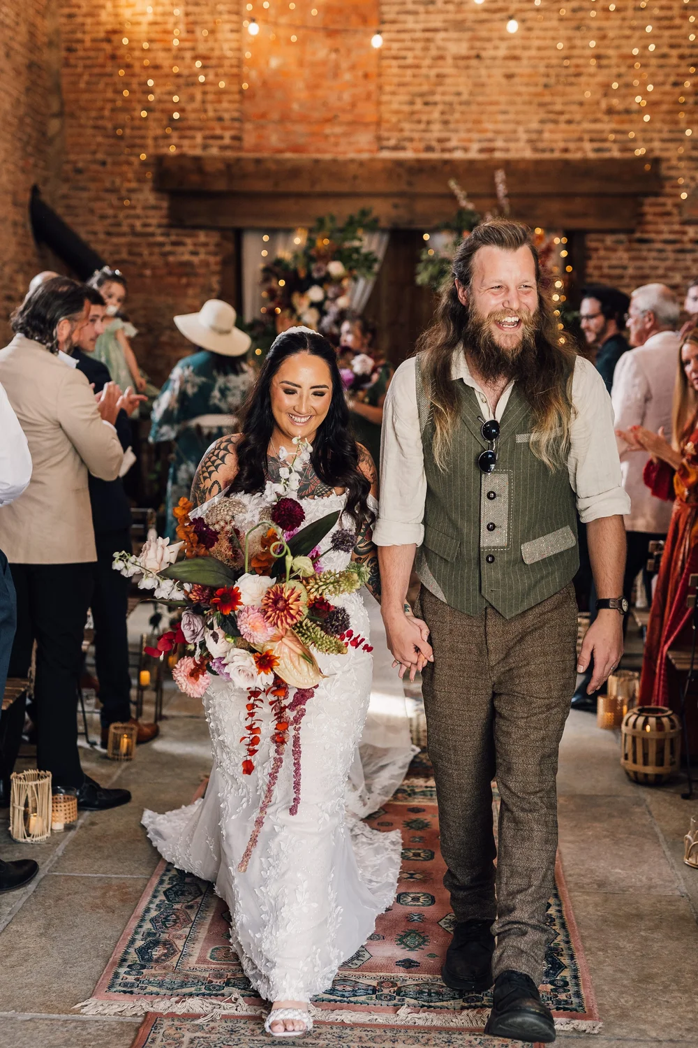 Bride and groom walking down aisle holding hands, surrounded by wedding guests in a rustic indoor setting with brick walls and string lights.