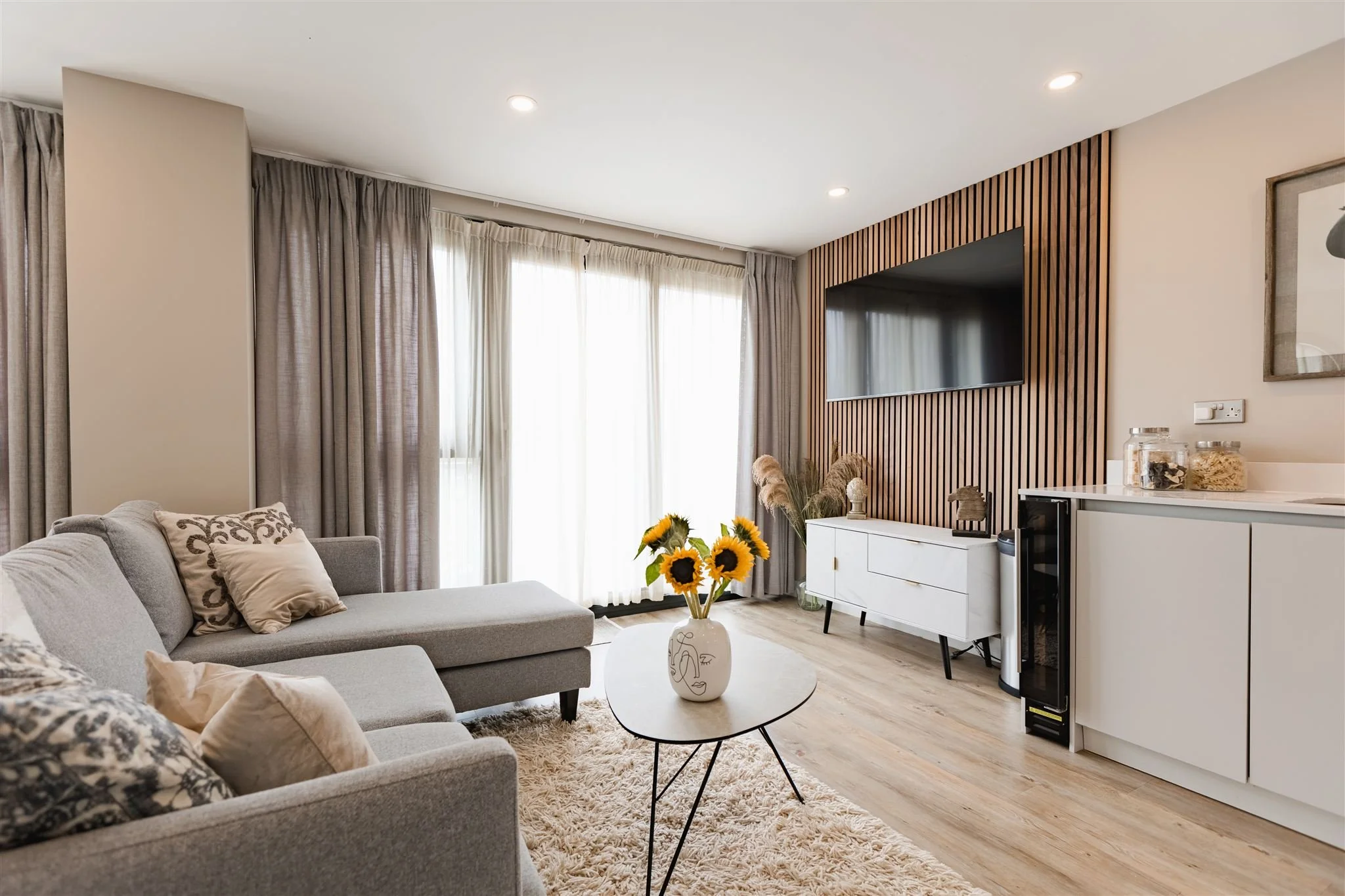 Modern living room with a gray sectional sofa, decorative pillows, a round coffee table with a sunflower vase, large windows with gray and white curtains, a wall-mounted TV on a wooden slat feature wall, and a white cabinet with jars and framed artwork.