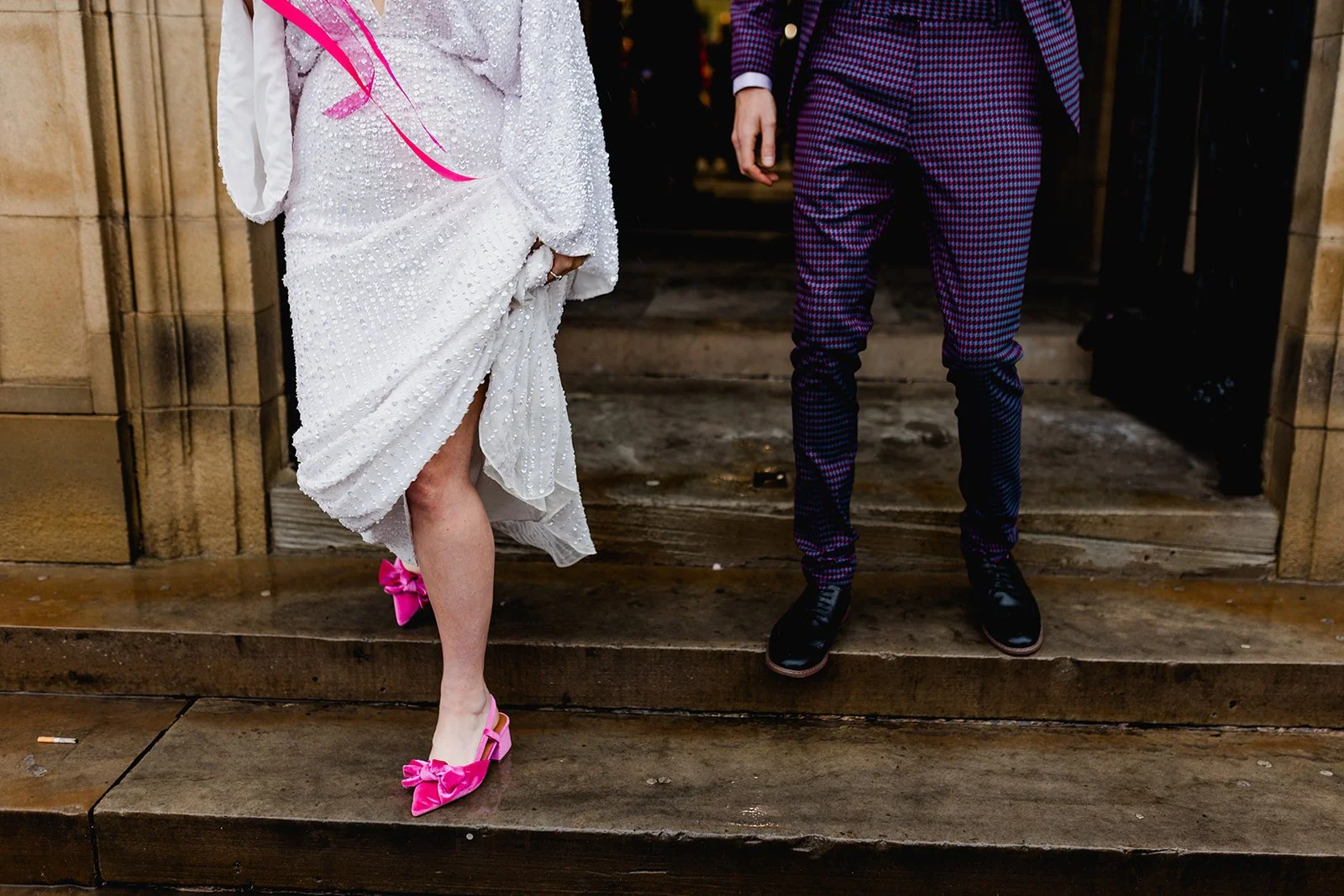 Partially visible people dressed in fancy attire standing on wet stone steps outside a building. One person wears a white dress with pearl embellishments and pink shoes and bows, and the other wears a purple patterned suit with black shoes.