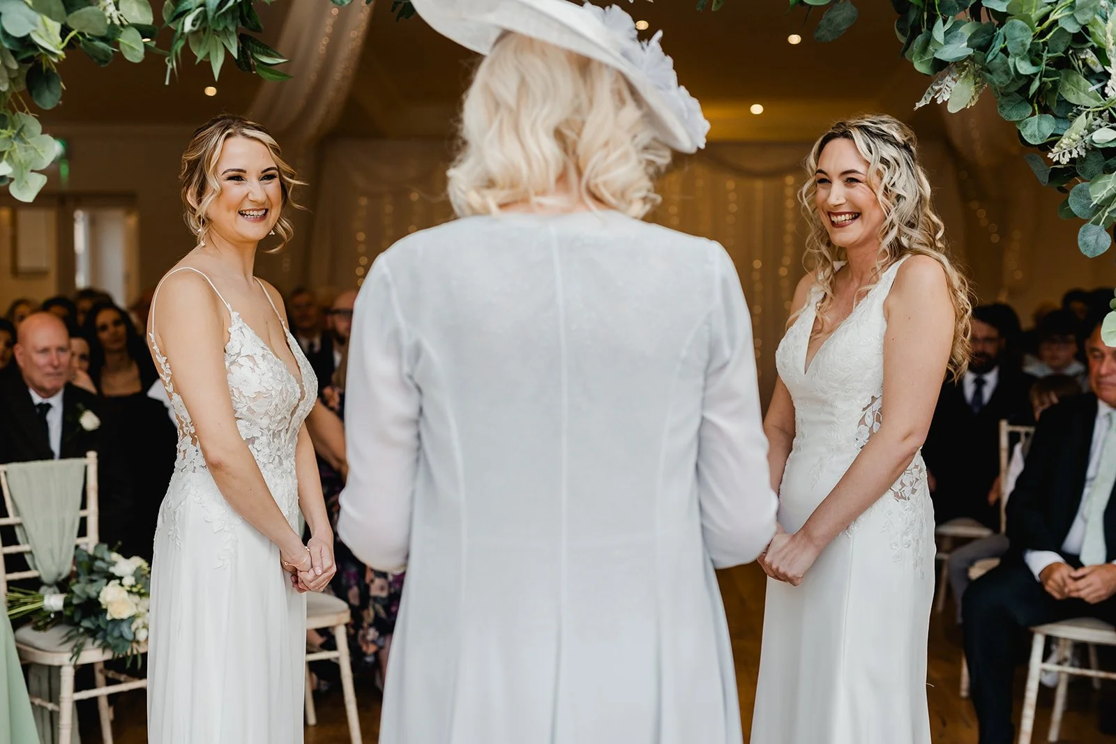 Two brides standing before an officiant during a wedding ceremony, smiling, with seated guests in the background.