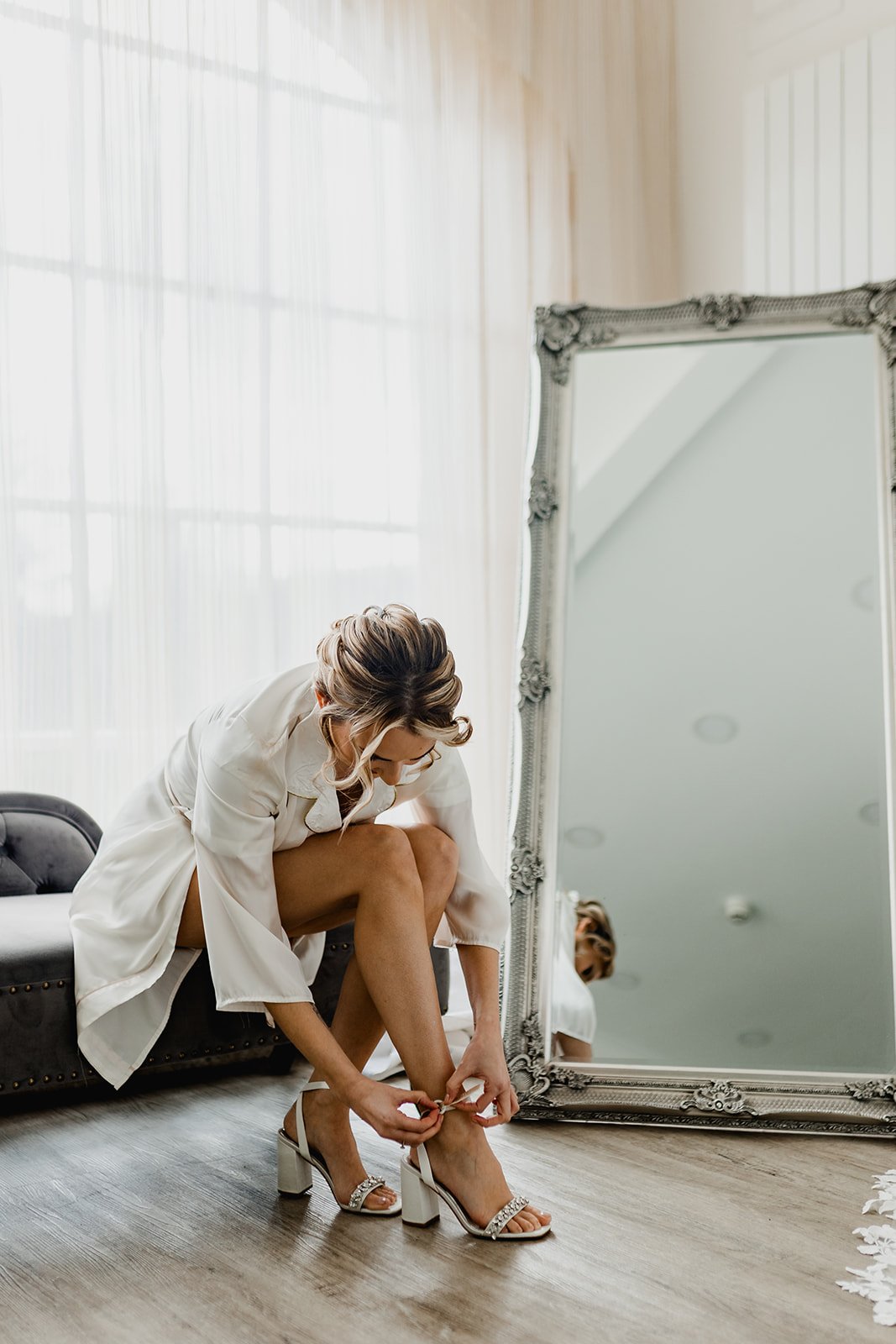 A Bride in a white robe putting on elegant high-heeled sandals while crouching in front of a large mirror in a bright room with sheer curtains.