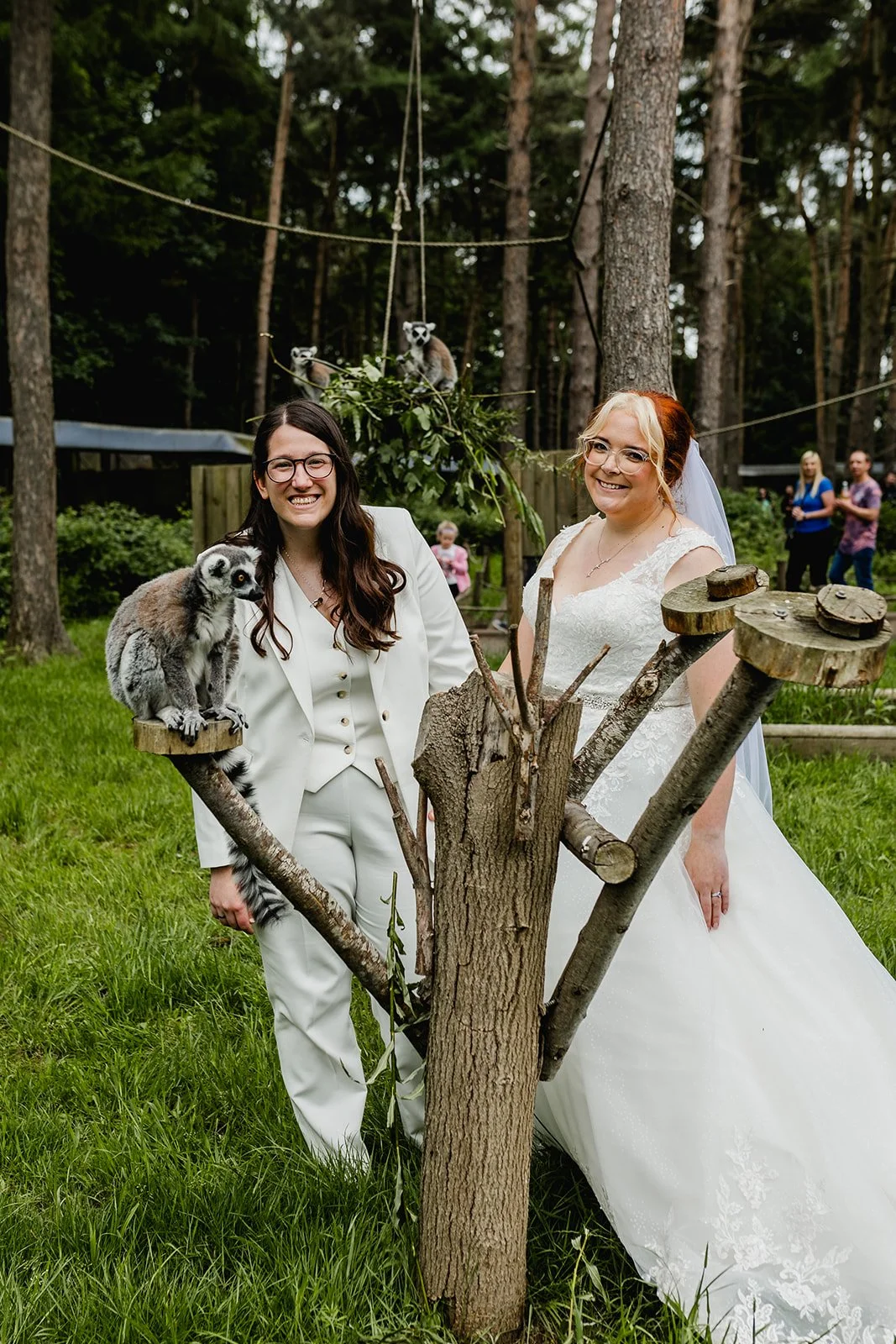 Two women, one in a white wedding dress and the other in a white suit, stand outdoors in a wooded area, smiling at the camera. They are surrounded by trees and grass, with a wooden structure and other people in the background. A lemur perches on a wo