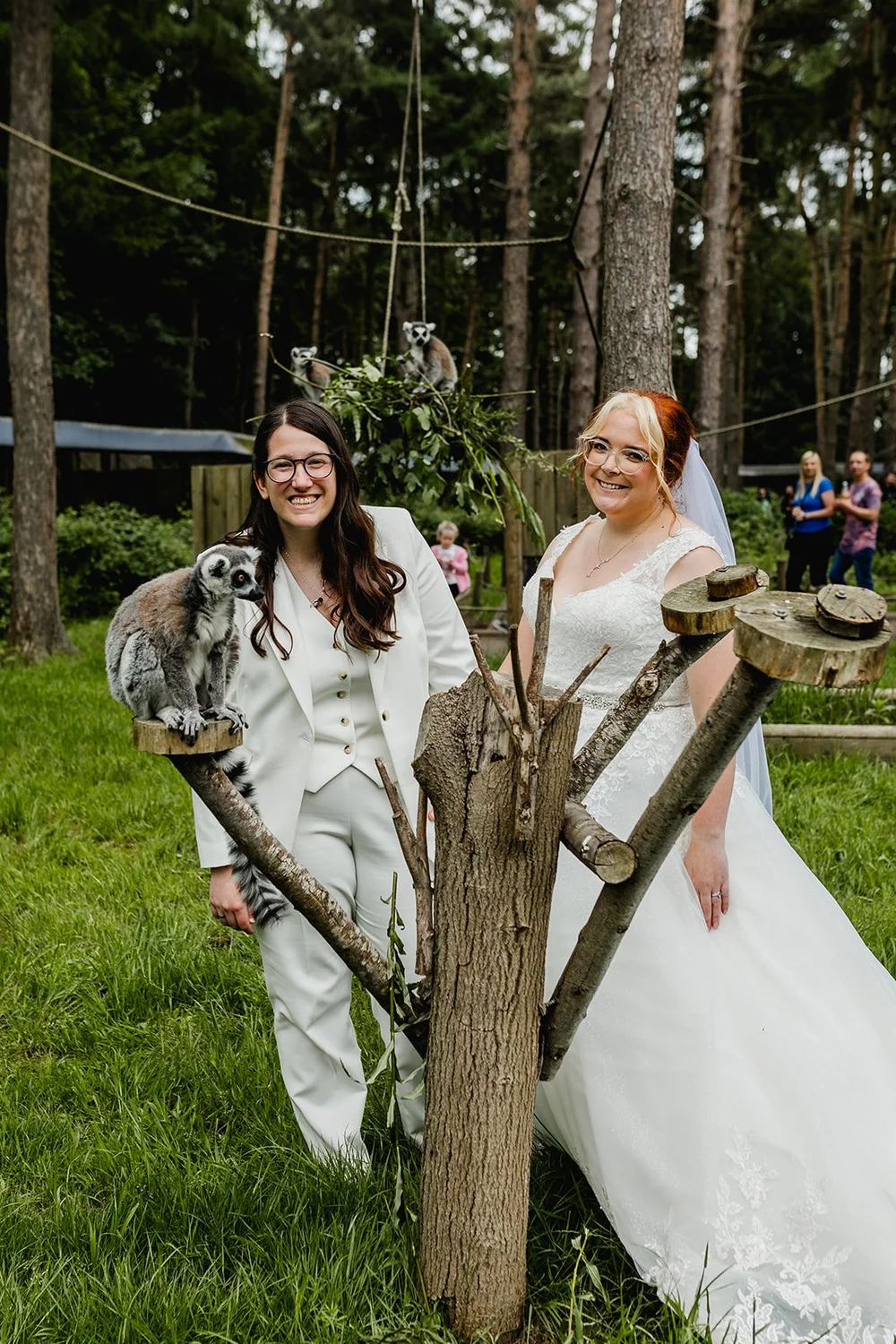 Two women, one in a white wedding dress and the other in a white suit, stand outdoors in a wooded area, smiling at the camera. They are surrounded by trees and grass, with a wooden structure and other people in the background. A lemur perches on a wo
