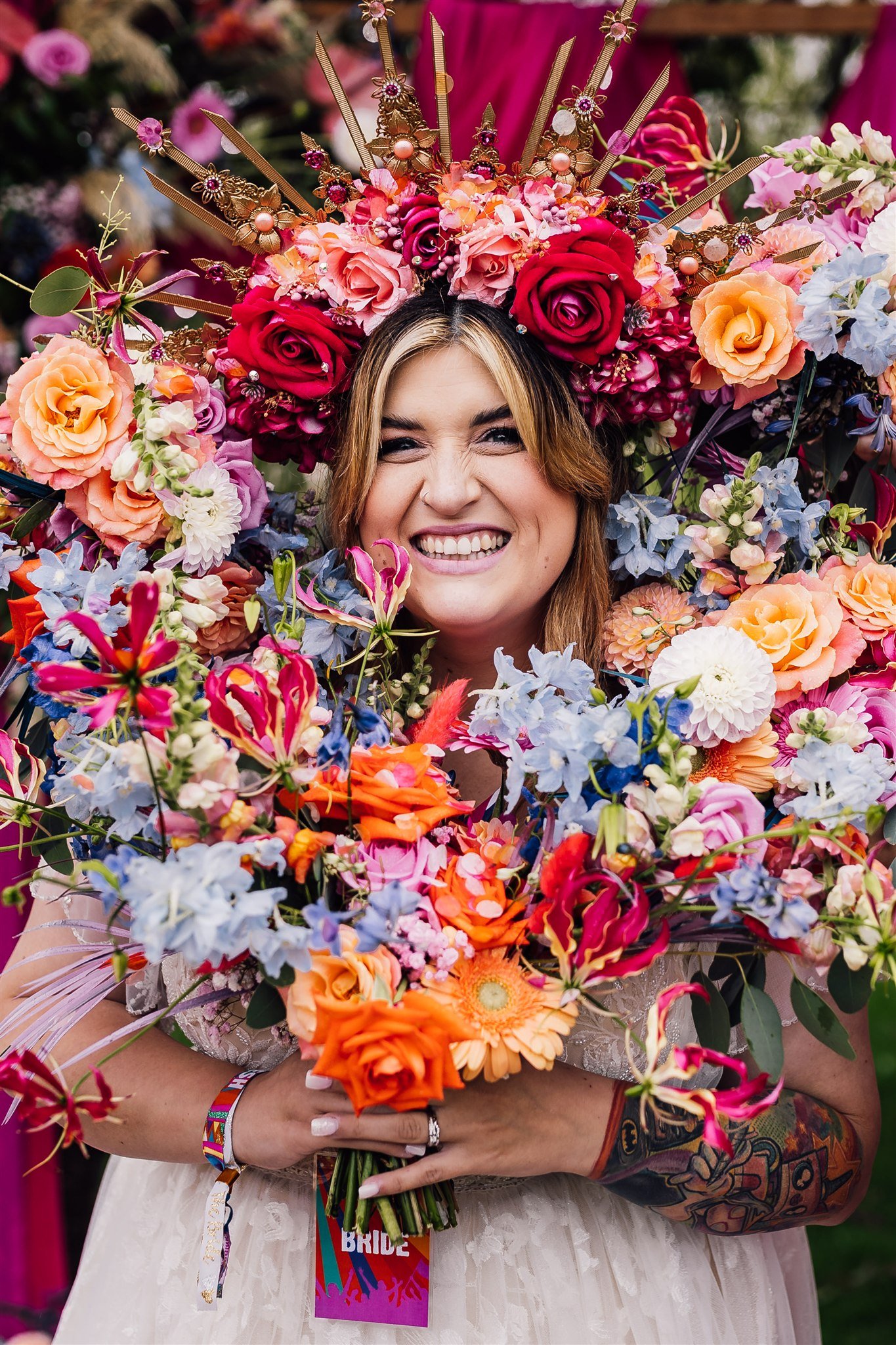 A smiling bride in a wedding dress with flowers surrounding her holding a boutique with a flower crown