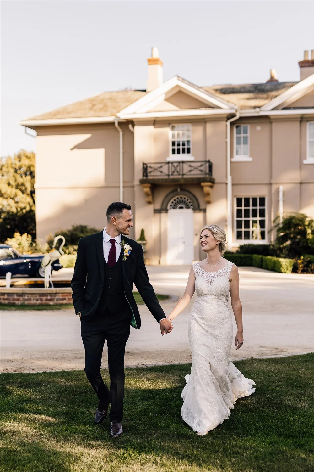 A bride and groom holding hands and walking outside a large beige house with a small balcony, greenery, and vintage cars in the background, on a sunny day.