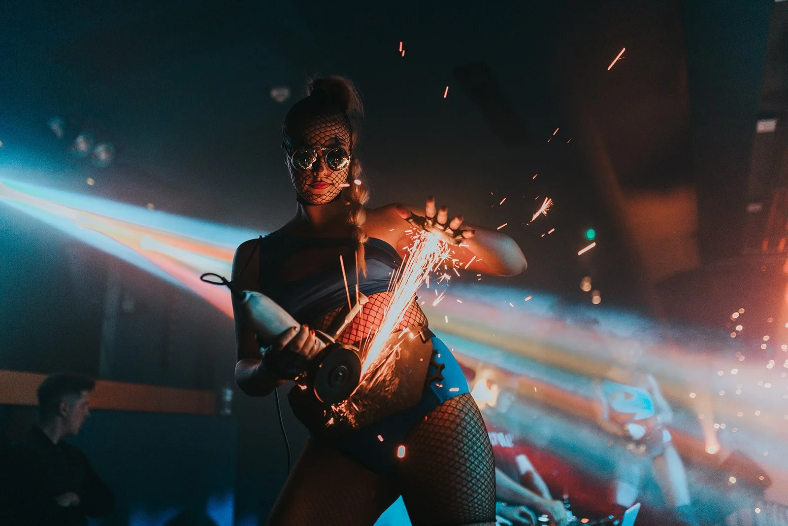 A woman in dark clothing and fishnet stockings holding a sparkler in a dark, colorful nightclub setting, wearing sunglasses and a mesh face covering.