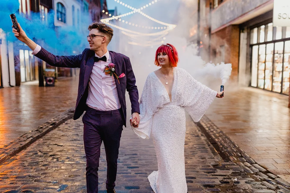 A couple dressed in wedding attire walks hand in hand down a cobblestone street at dusk, holding smoke canisters emitting white smoke, with string lights overhead and storefronts on both sides.