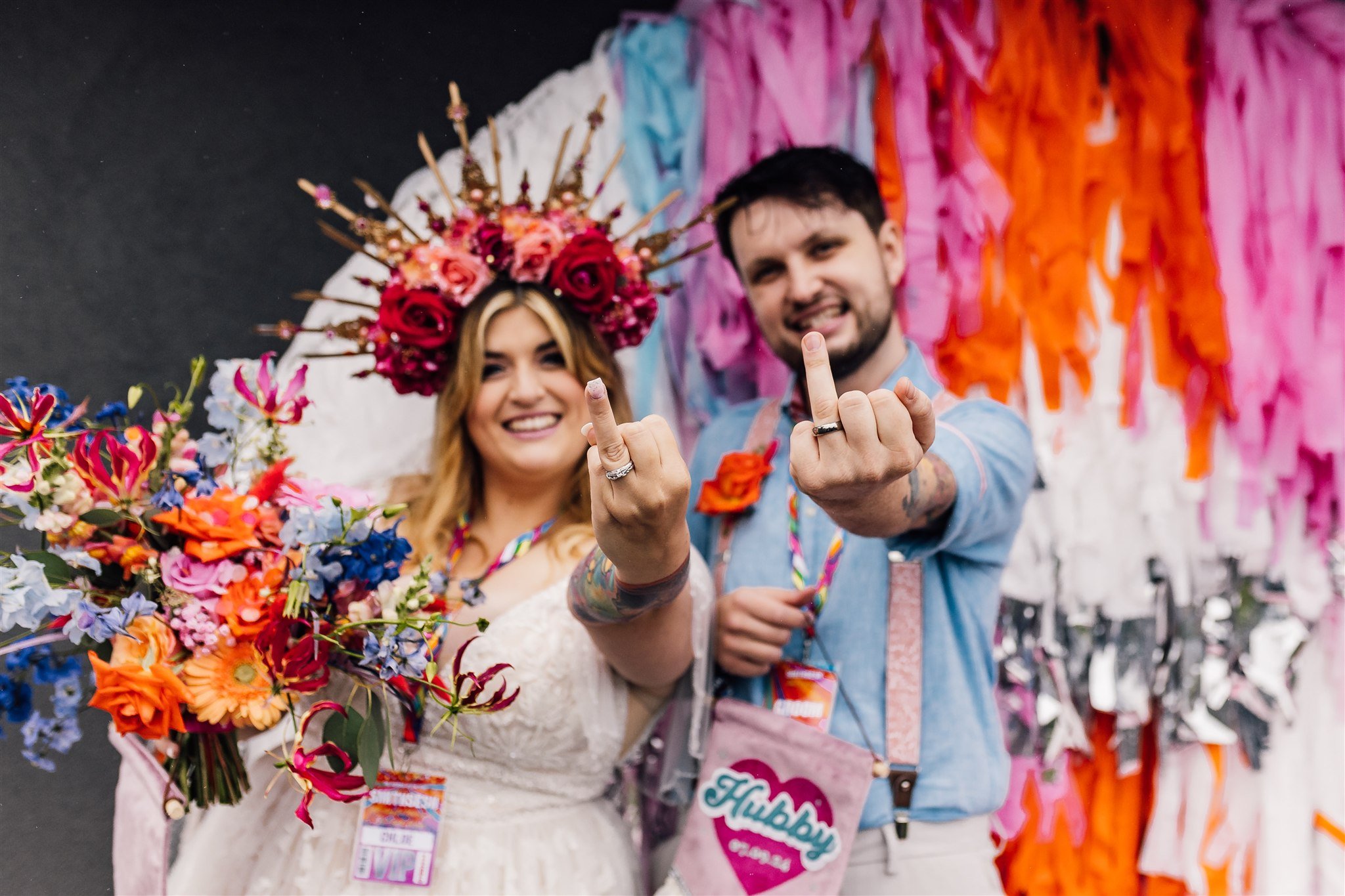 A smiling bride wearing a floral crown and holding a bouquet, and a smiling groom showing their middle fingers, at a colorful wedding or festival, surrounded by pink, orange, and purple decorations.