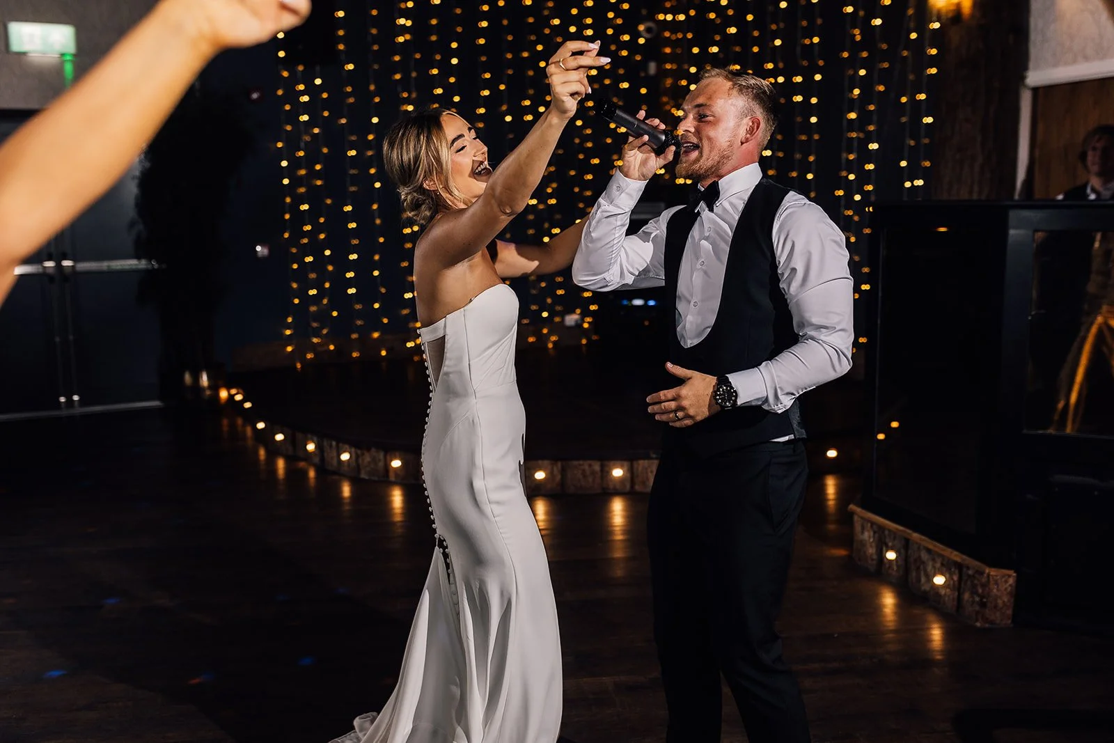 A bride and groom singing together and dancing at their wedding reception, with a dark background and warm string lights.