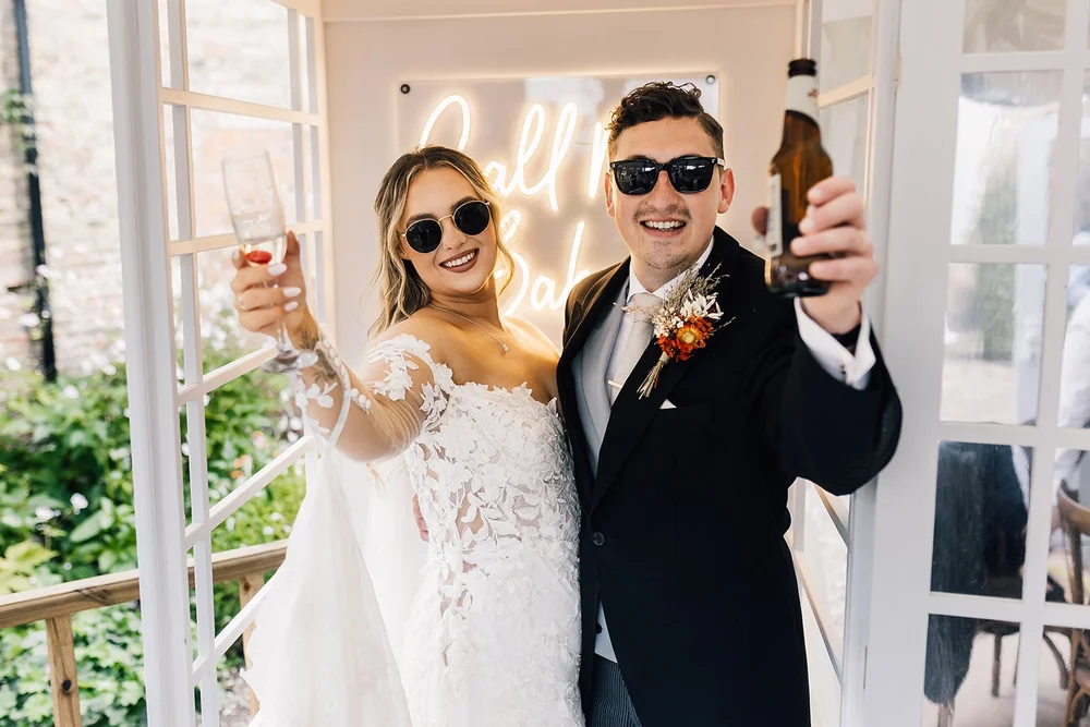 A happy bride and groom celebrating at their wedding, wearing sunglasses, holding drinks, and smiling at the camera inside a decorated space.