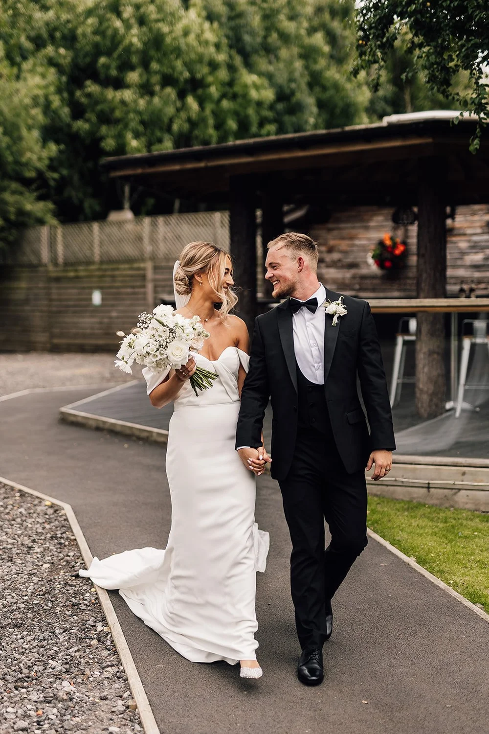 A bride and groom walk hand in hand outdoors, smiling at each other, with the bride holding a bouquet of white flowers.