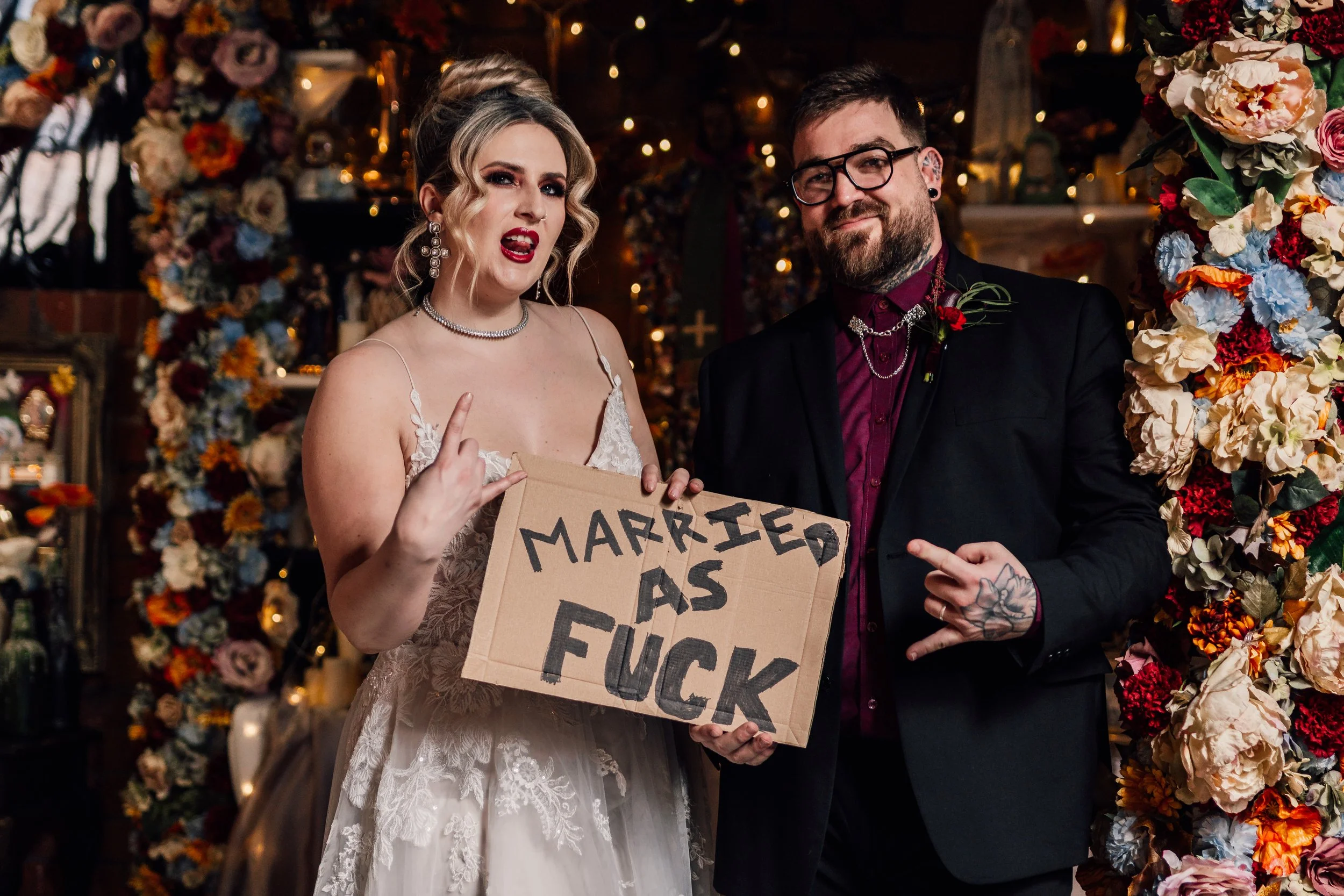 Alternative wedding couple holding sign “married as fuck” at Church Temple of Fun Sheffield with colourful florals and candlelit backdrop