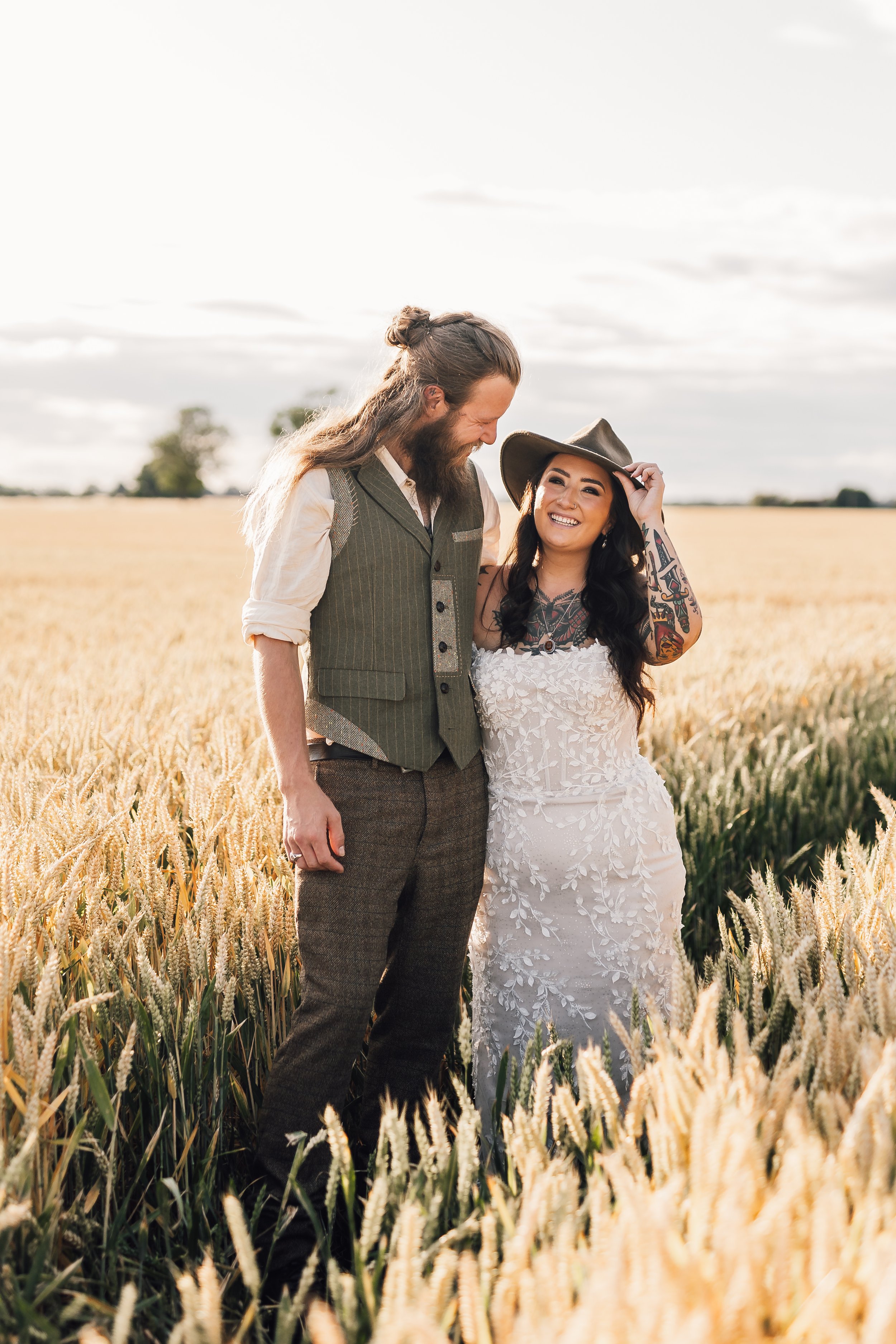 A happy couple standing in a wheat field, smiling at each other. The woman wears a white lace dress and a wide-brimmed hat, while the man wears a vest and dress pants. The scene is outdoors with a bright sky and some clouds.