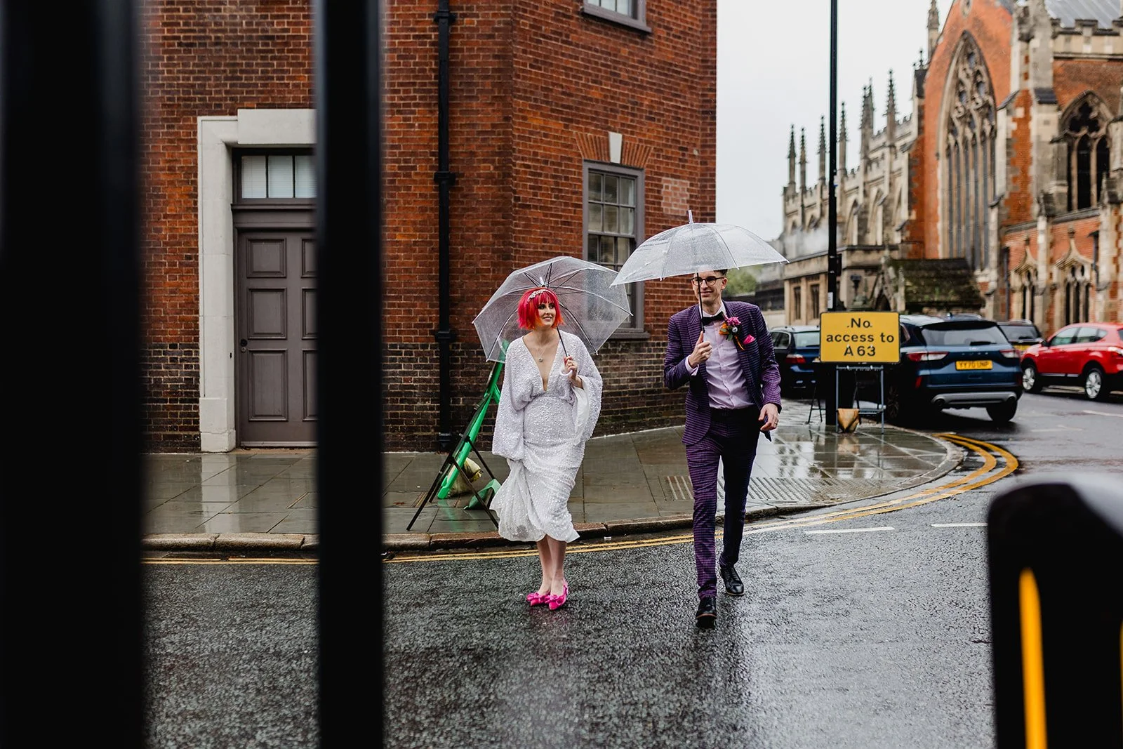 A man and woman walking on a rainy city street with umbrellas. The woman has bright pink hair and is wearing a white dress and pink shoes, while the man is wearing a purple checkered suit.