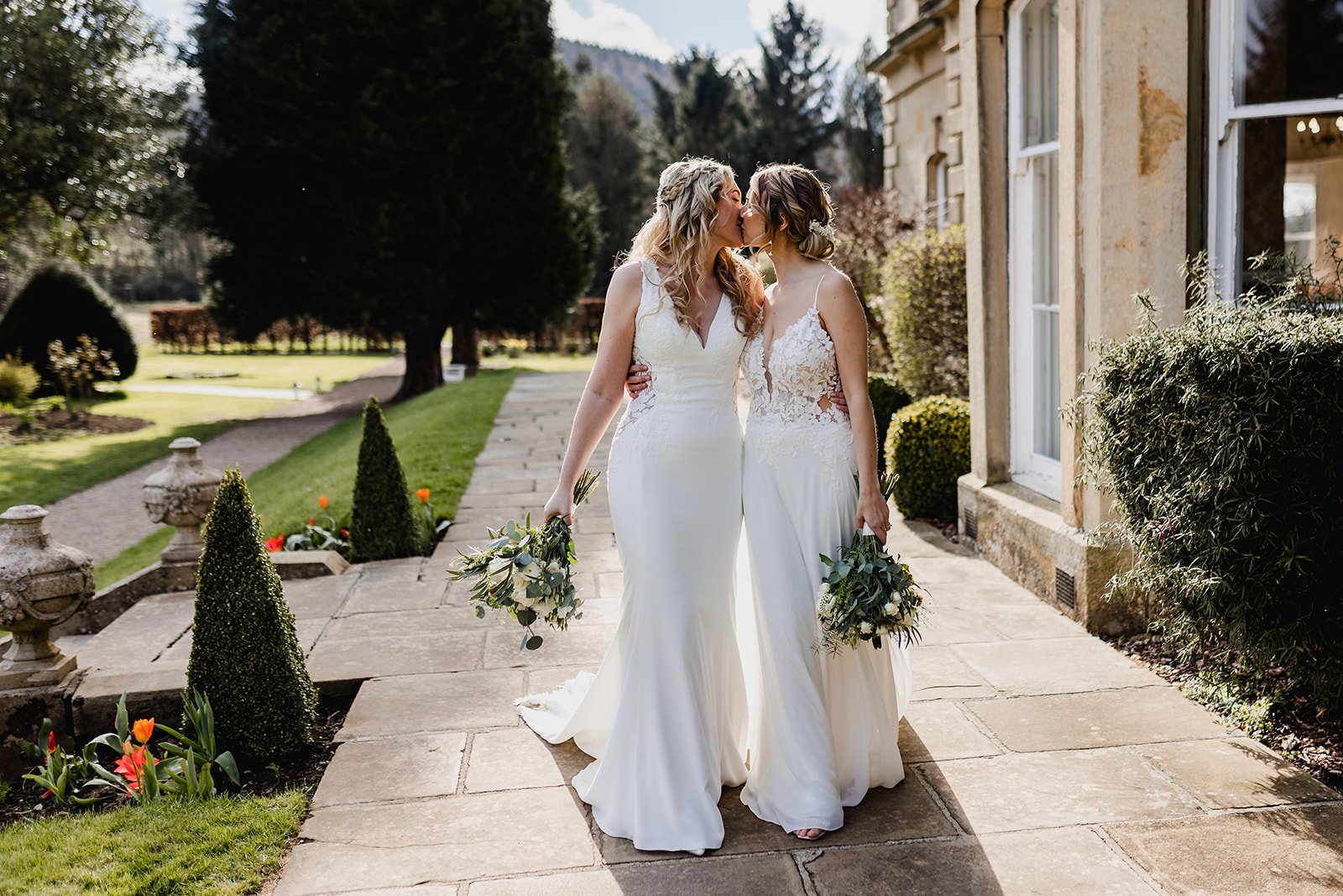 Two Brides in wedding dresses sharing a kiss outdoors on a stone pathway, holding bouquets, surrounded by greenery and a historic building.