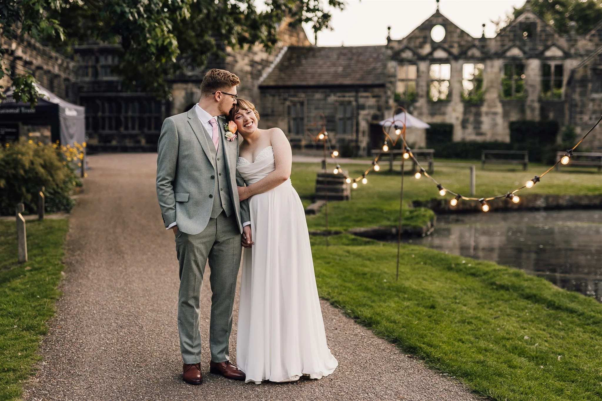 Bride and groom standing by the lake at East Riddlesden Hall with festoon lights and historic building behind