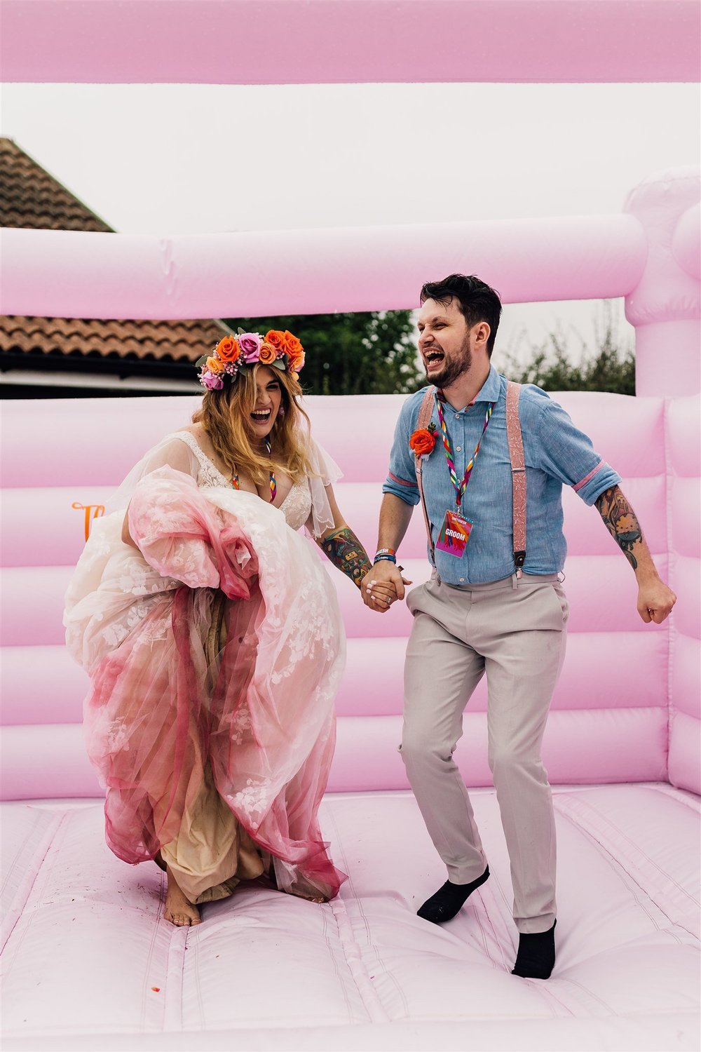 A Bride and Groom are on a pink bouncy castle, holding hands, laughing, and celebrating. The woman is wearing a floral crown and a flowing dress, while the man is dressed casually suspenders, and a flower boutonniere.