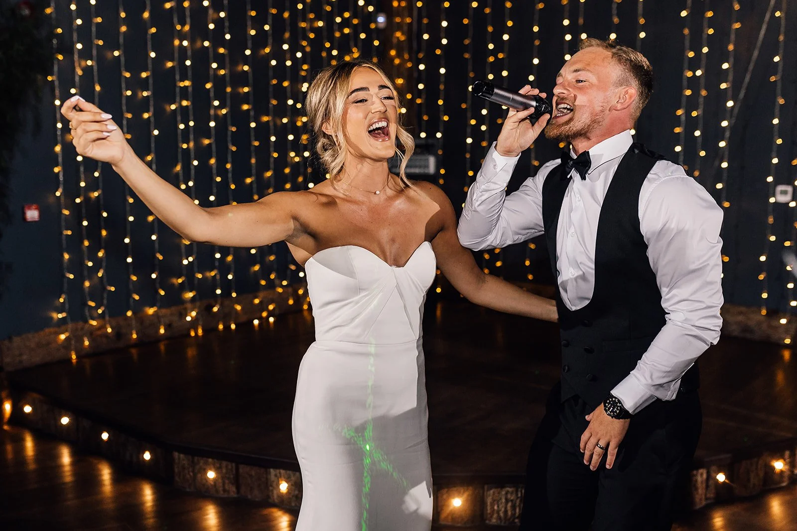 A bride and groom singing and dancing at their wedding reception with string lights in the background.