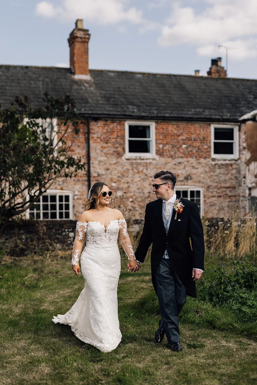 A bride and groom walking hand-in-hand outdoors in front of an old brick building during a wedding.
